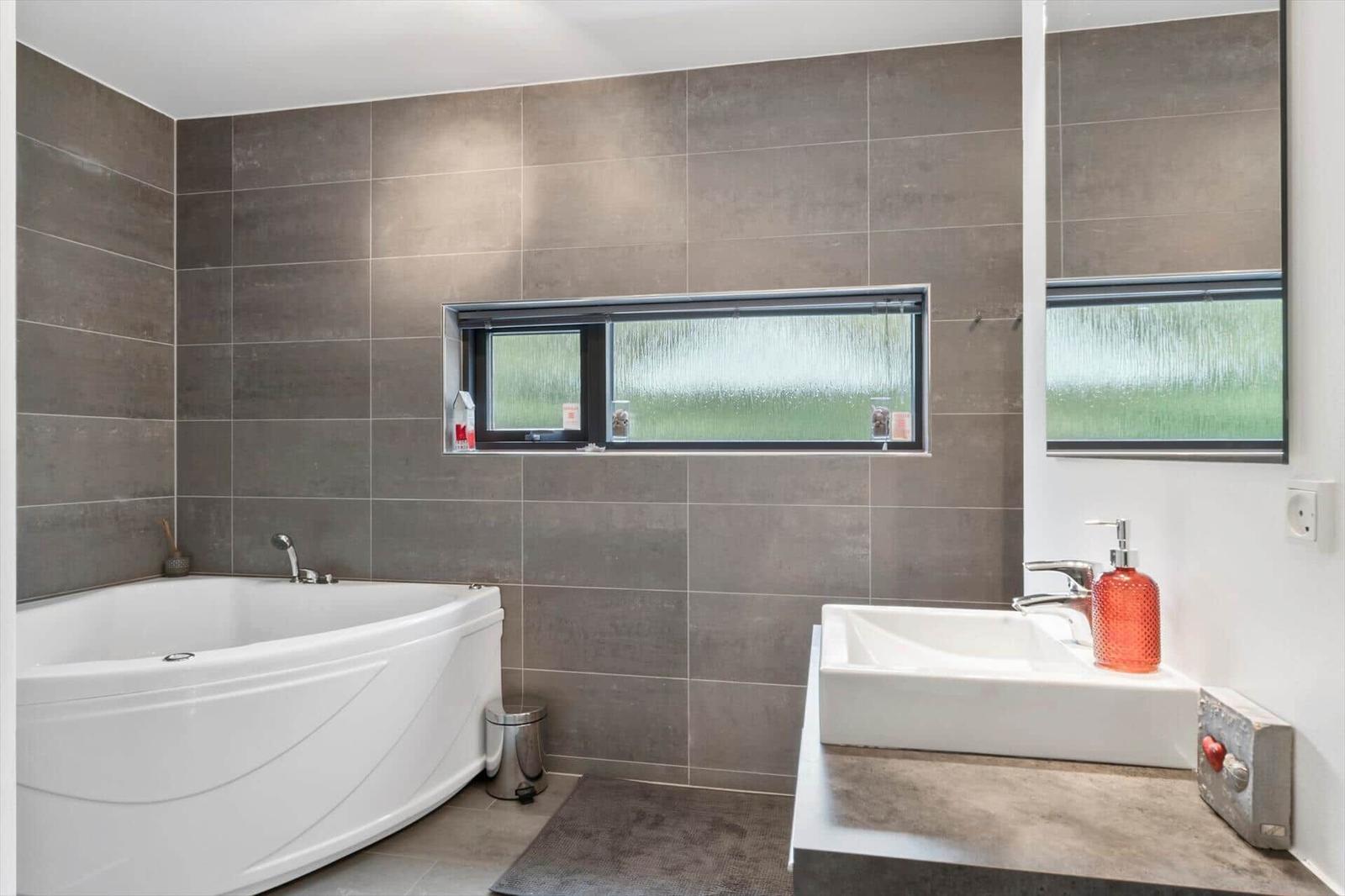 Bathroom with white corner tub, grey tiles and a vessel sink.
