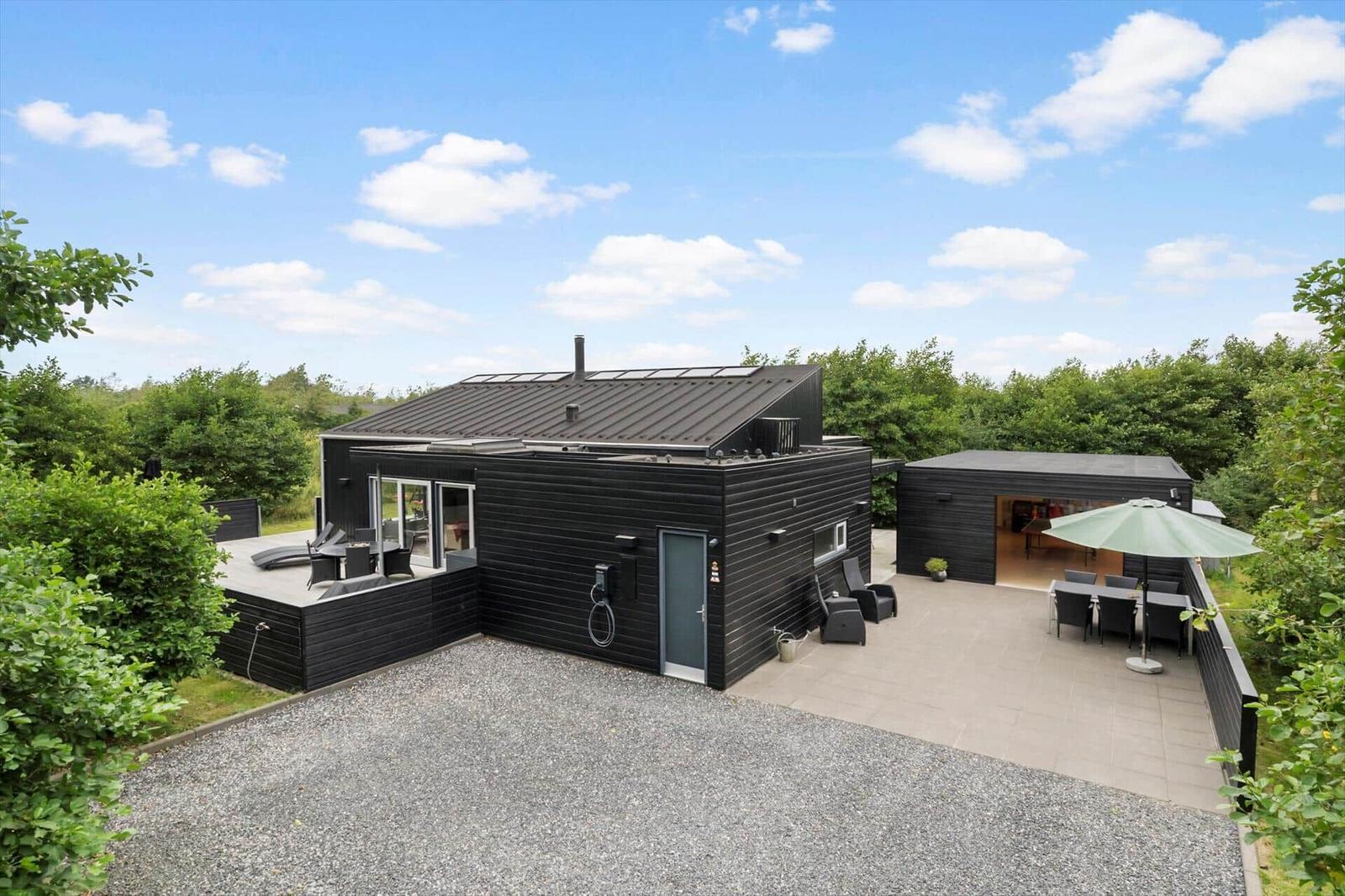 Black wooden house with gravel, terrace, green parasol and an outbuilding.