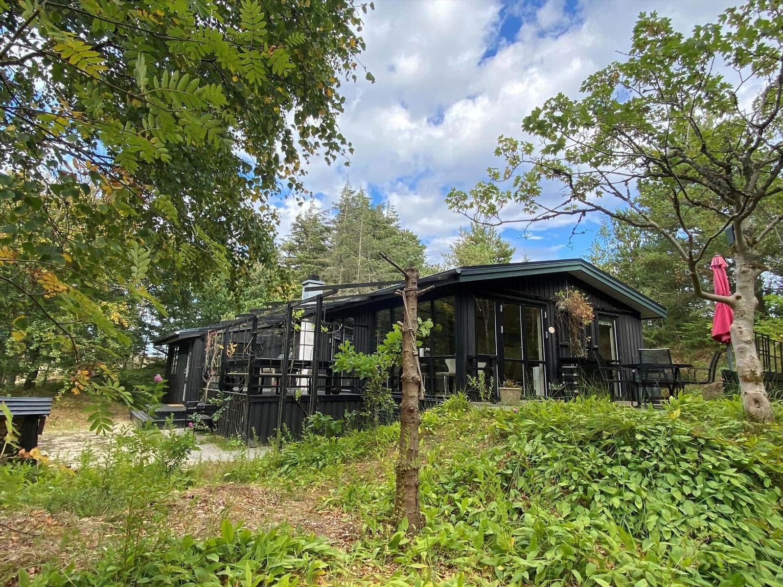 Black wooden house with glass front, wooden terrace, seating and white chimney.