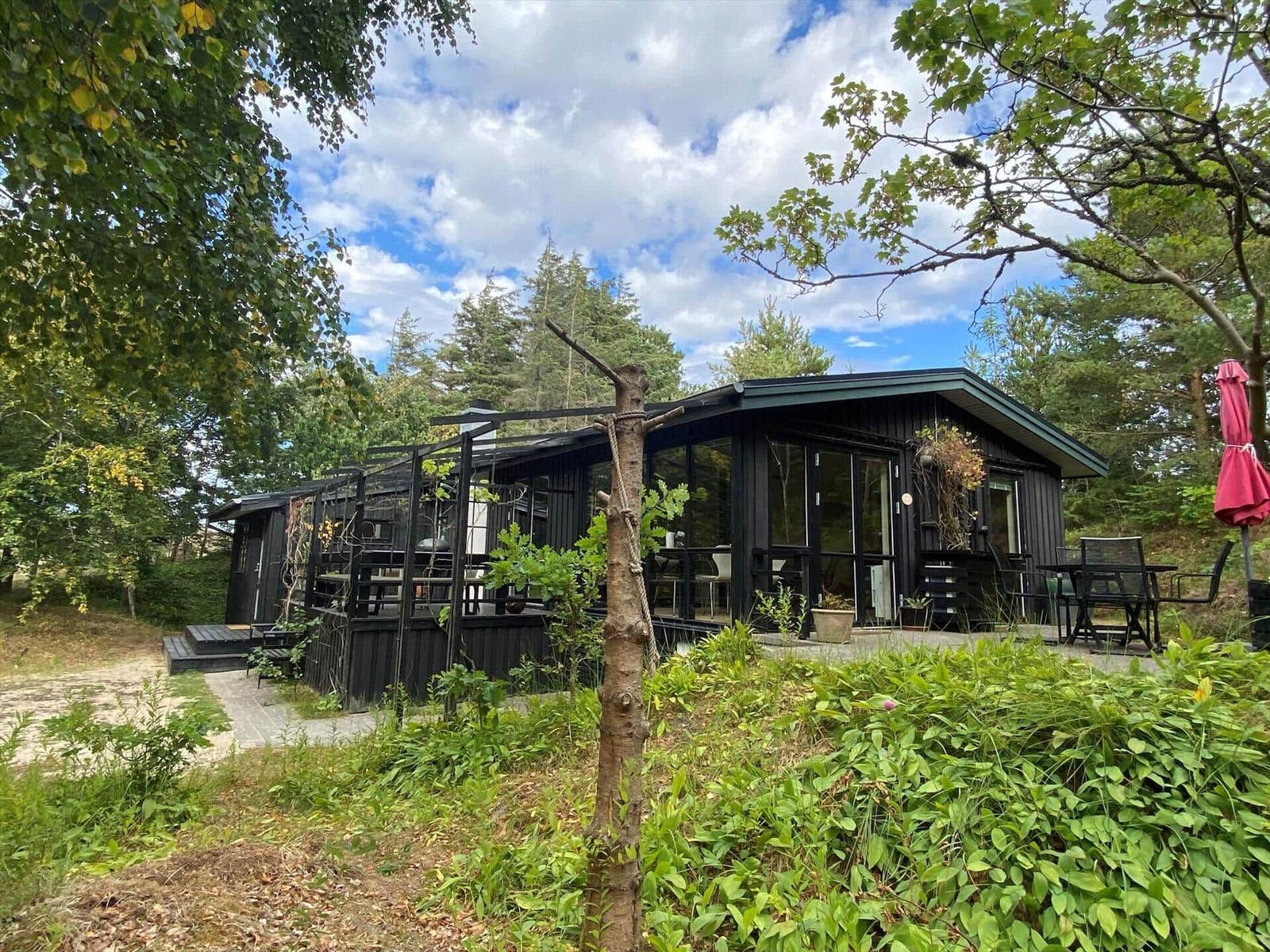 Black wooden house with wooden terrace, pergola, seating and red parasol.