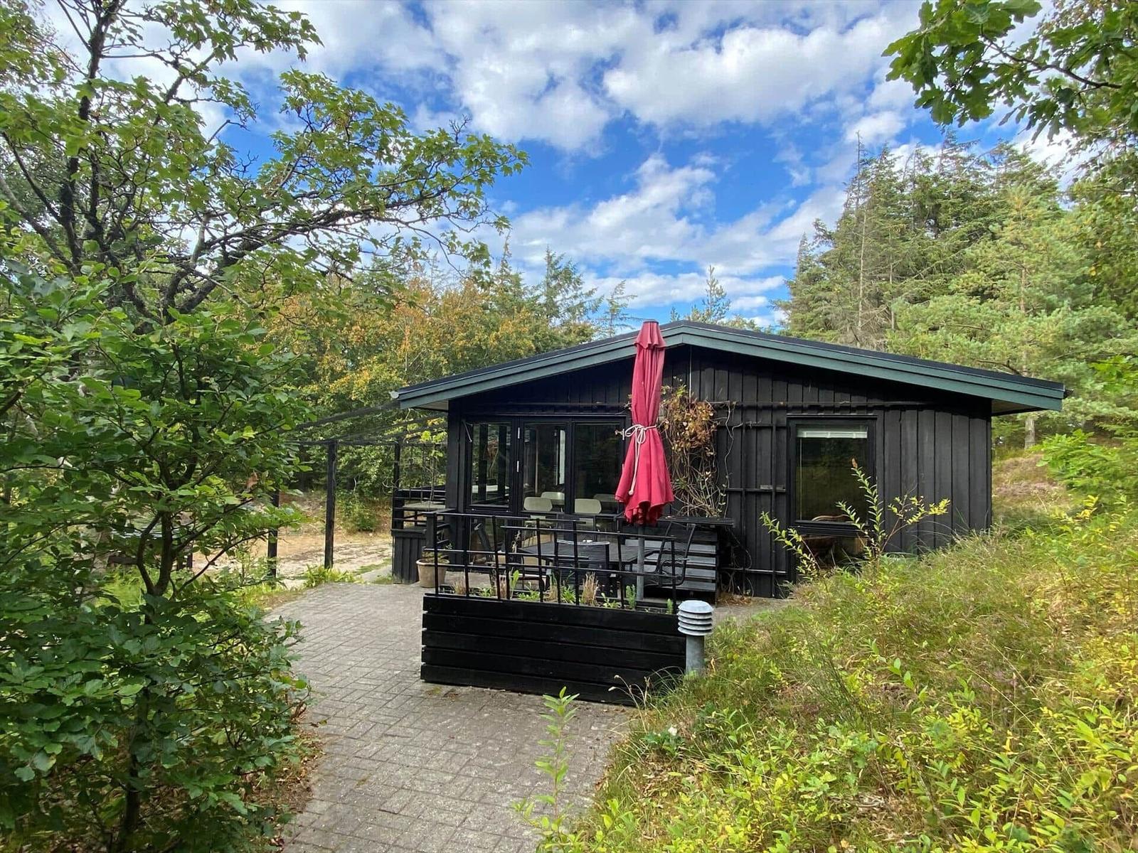 Black wooden house with terrace, seating, closed red parasol and trees.