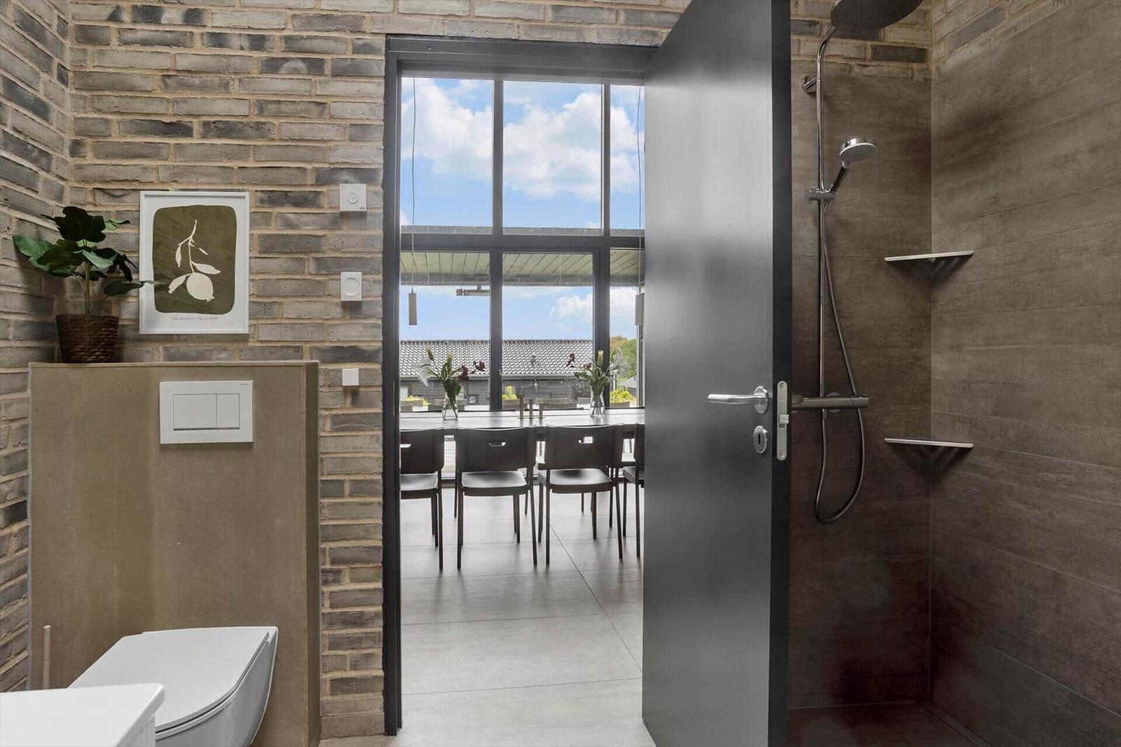 Bathroom with toilet, walk-in shower and view of dining table by windows.
