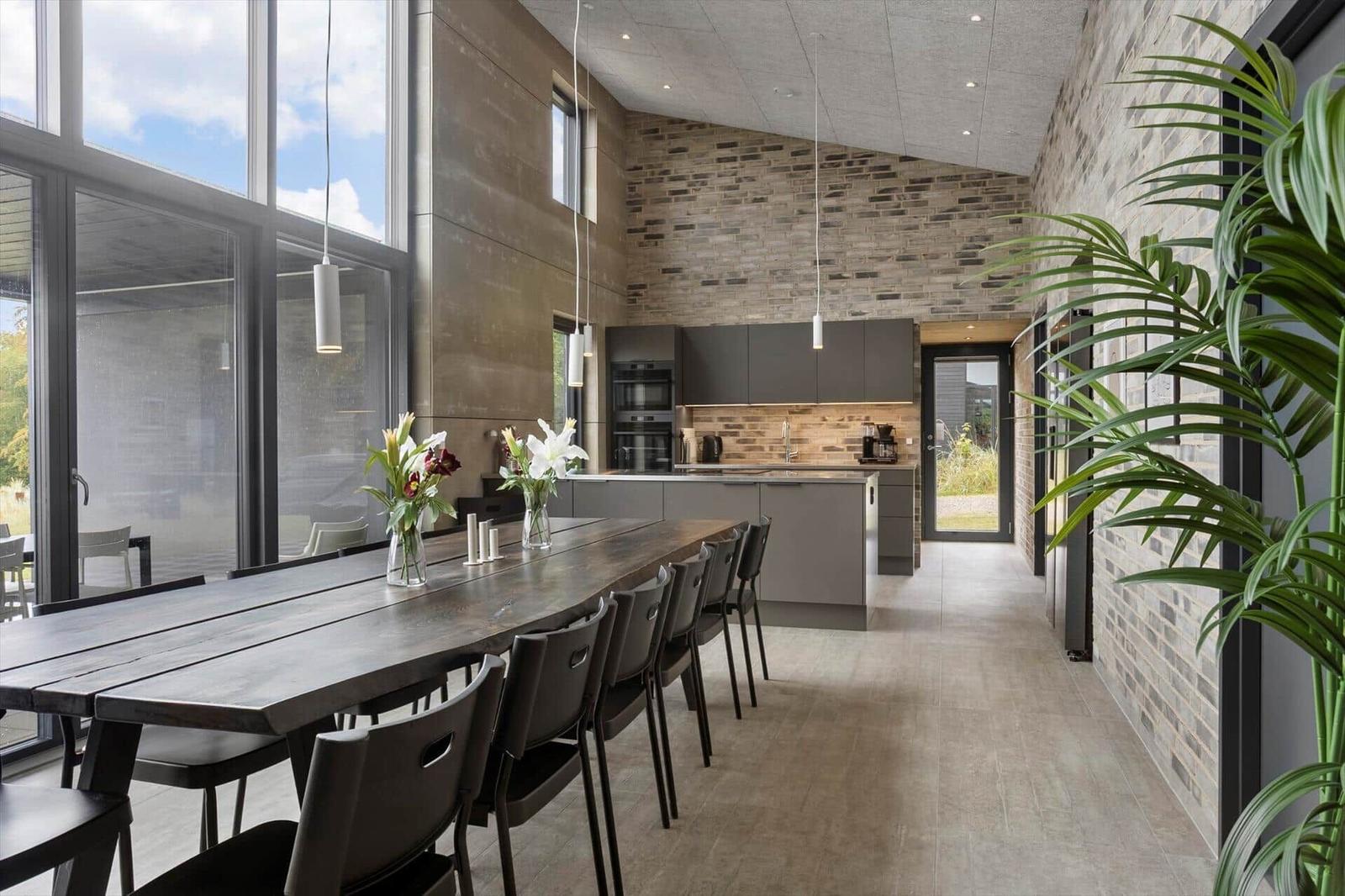 Open living space with long dining table, dark kitchen and glass front.