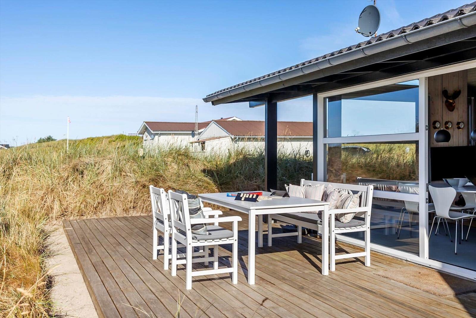Wooden terrace with white table, chairs and bench by dune grass and nearby houses.