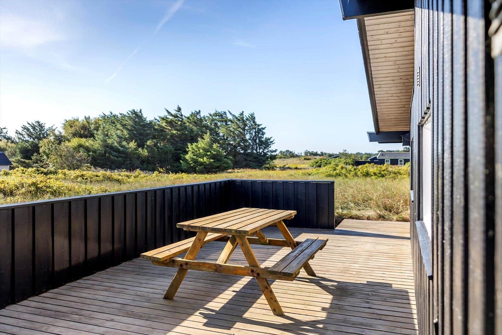 Wooden terrace with picnic table, black facade and a view of meadow and trees.