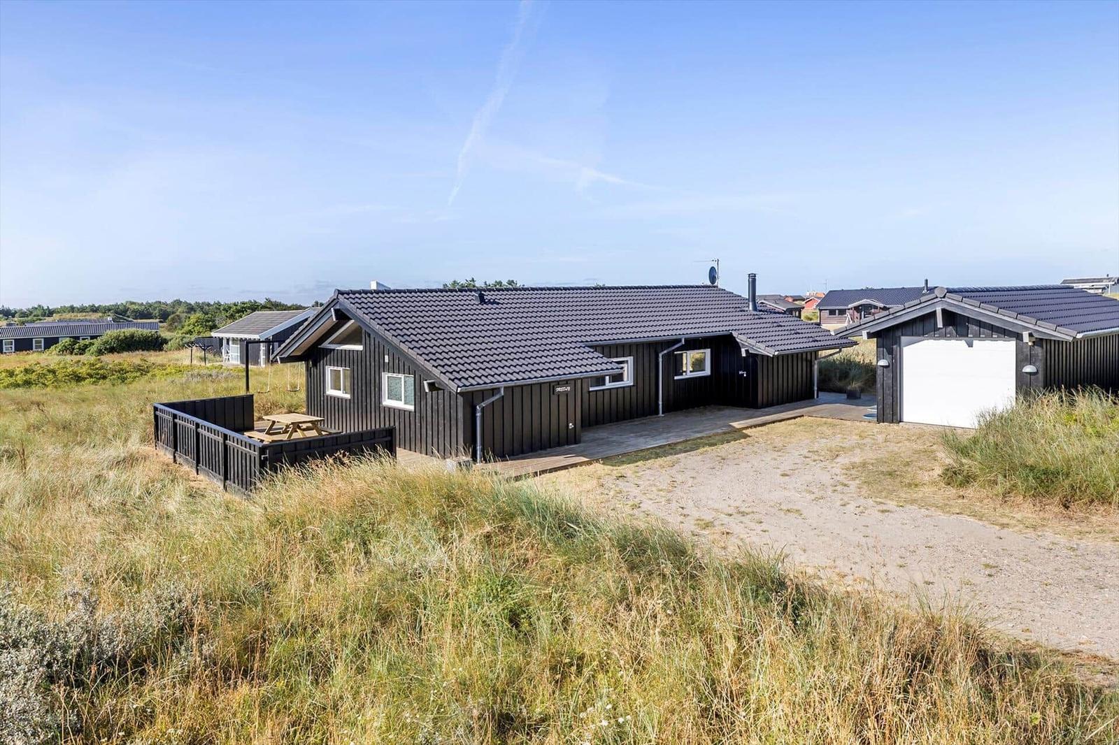 Black wooden house with terrace, garage with white door and dune grass under blue sky.