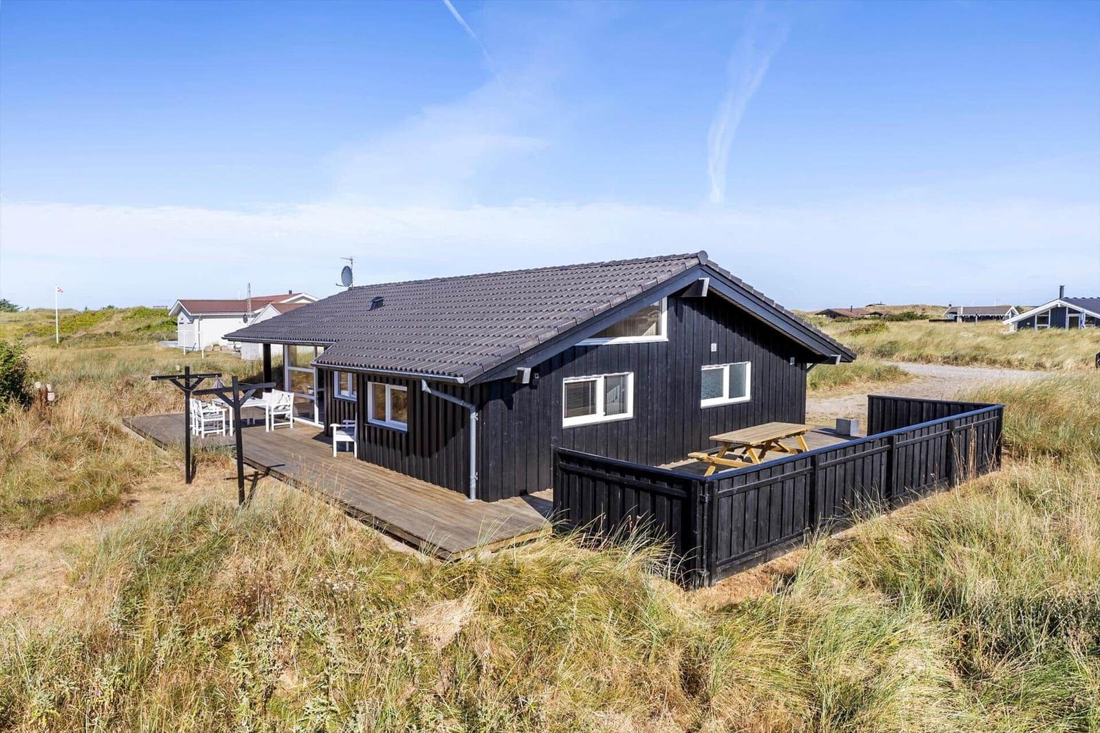 Black wooden house with white windows, wooden terrace and dune grass.