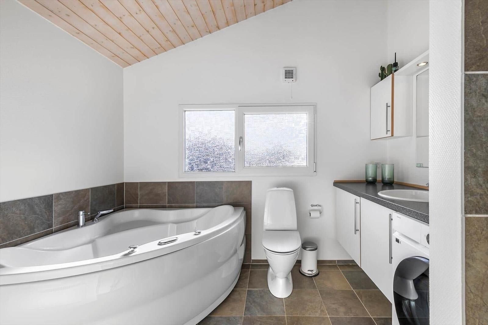 Bathroom with corner tub, toilet, vanity and washing machine under a sloped ceiling.