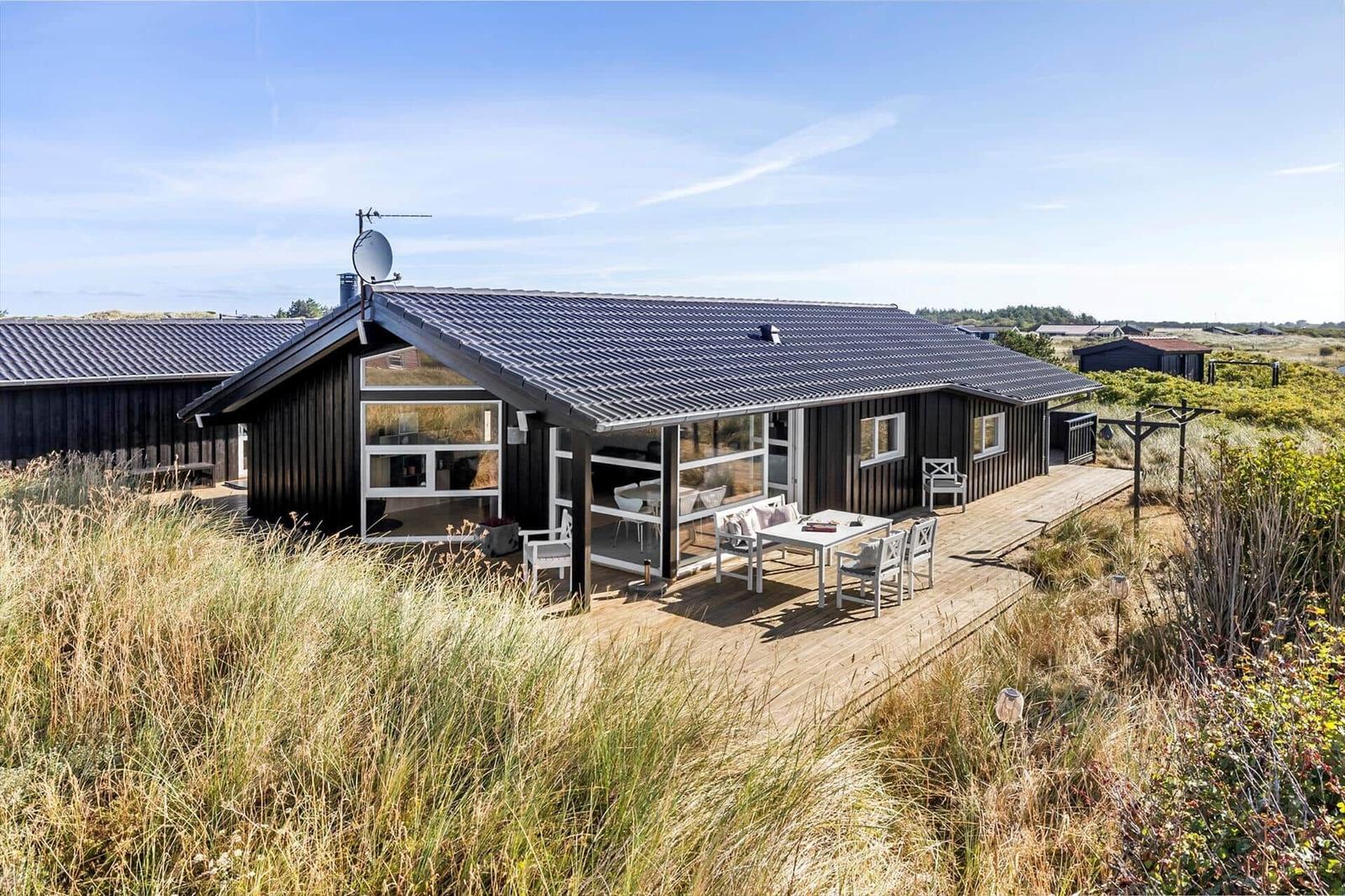 Black wooden house with large windows, wooden terrace and white seating in dune grass.