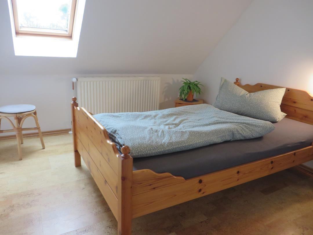 Holzbett mit gebogenem Kopfteil, Hocker und Pflanze unter Dachfenster.