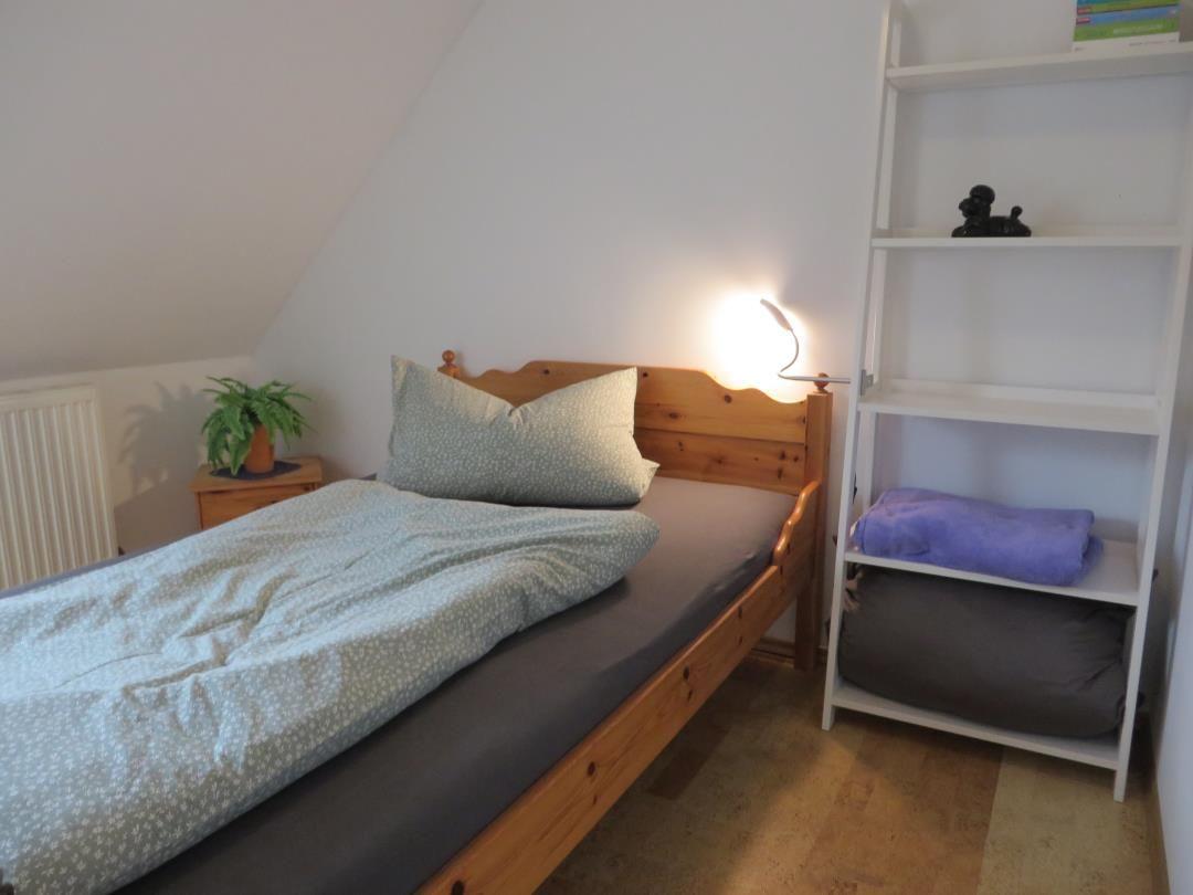Einzelbett aus Holz mit Leselampe, weißem Leiterregal und Pflanze.