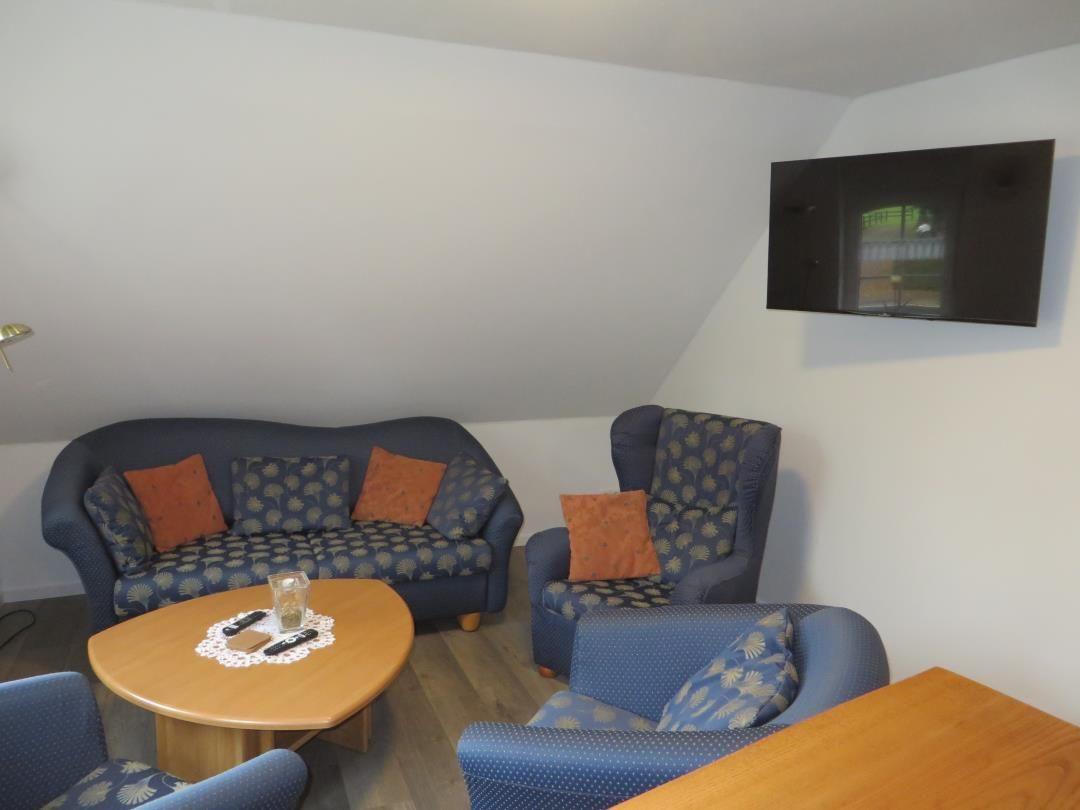 Wohnzimmer mit blau gemusterter Couch, zwei Sesseln, Holztisch und Fernseher.