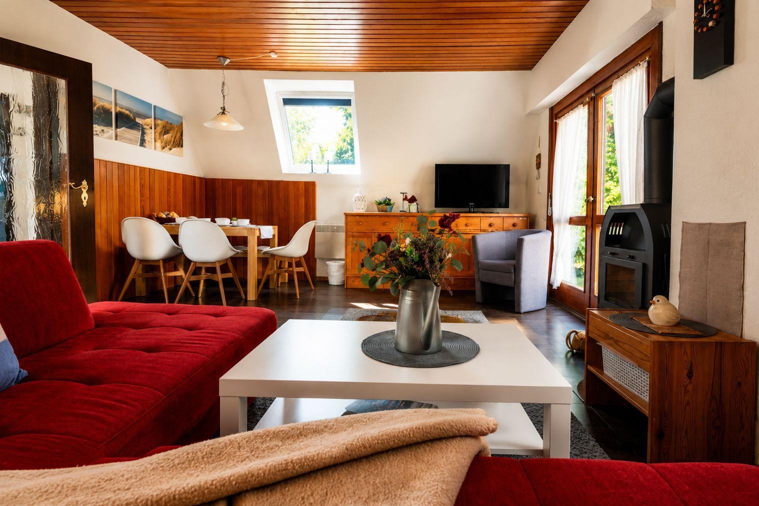 Wohnzimmer mit rotem Sofa, Esstisch, Fernseher und Kaminofen unter Holzdecke.