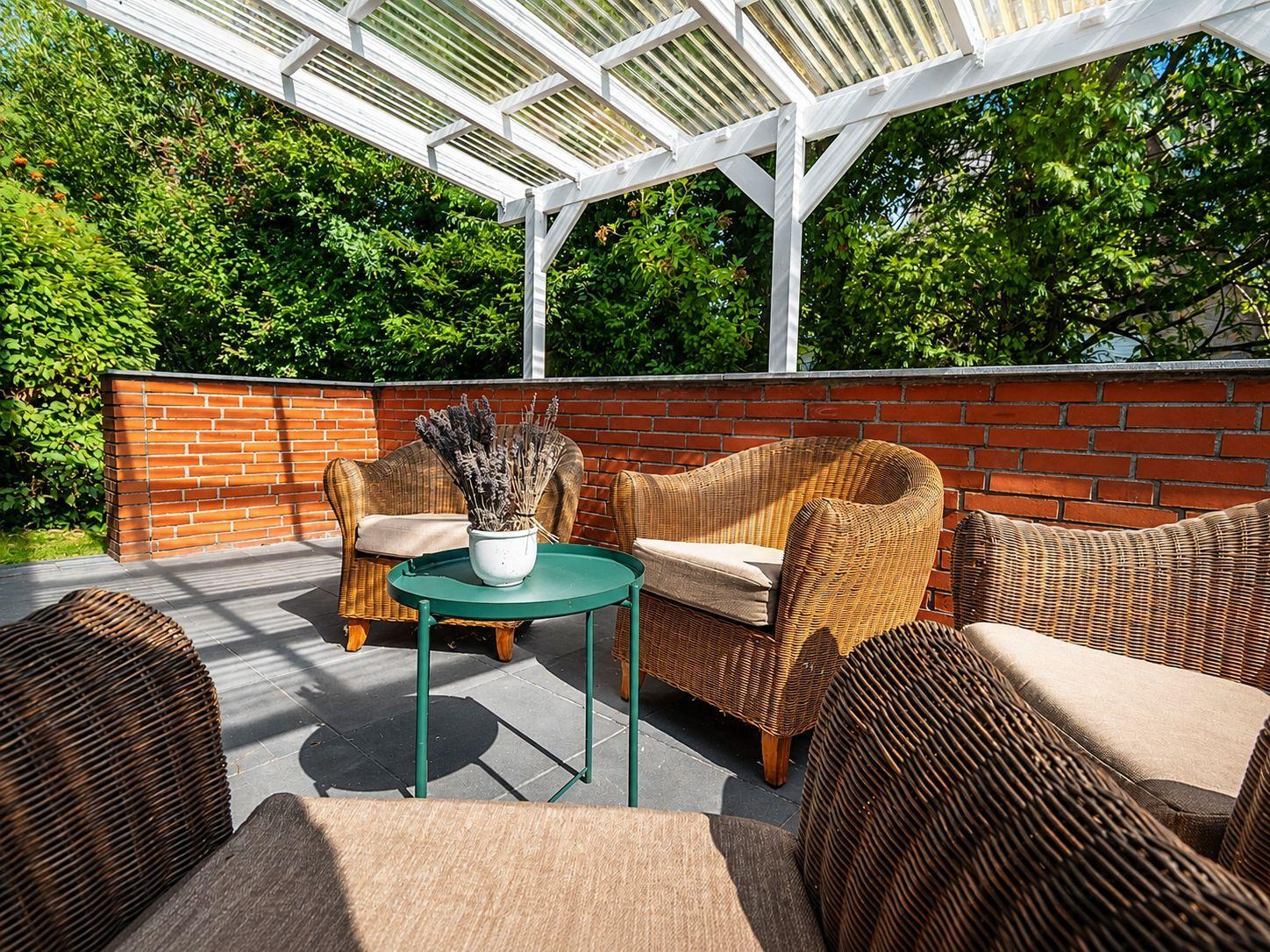Überdachte Terrasse mit Korbsesseln, rundem Tisch und Pergola vor Hecke.