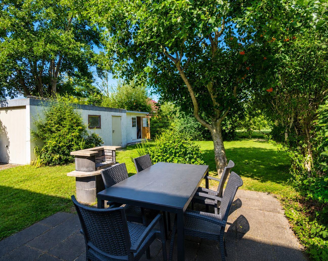 Gartenterrasse mit Esstisch, sechs Stühlen und Steingrill vor Rasen.