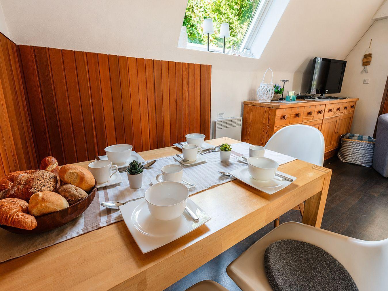 Gedeckter Holztisch mit weißen Stühlen, Holzbank und Sideboard mit Fernseher.