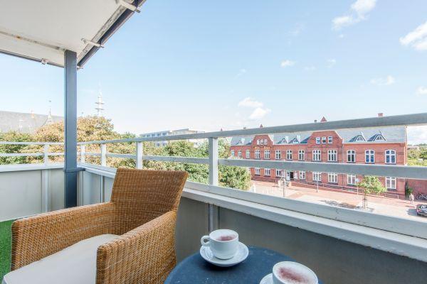 Balkon mit Korbsessel, rundem Tisch und Tassen vor Backsteinhaus.