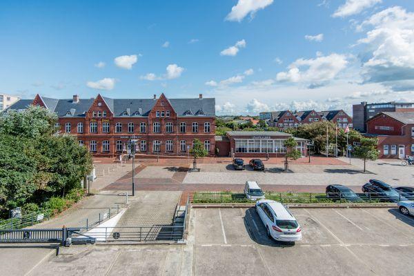 Blick auf einen Platz mit Backsteinhaus, Flachdachbau und parkenden Autos.