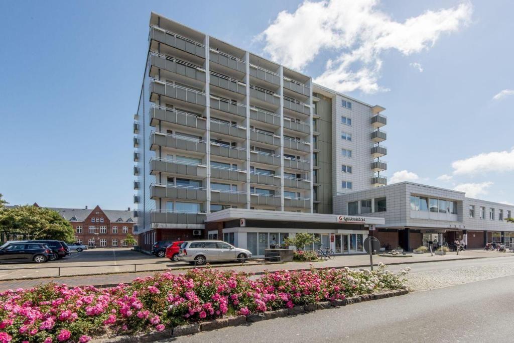 Mehrstöckiges Haus mit Balkonen, Rosenbeet und parkenden Autos davor.