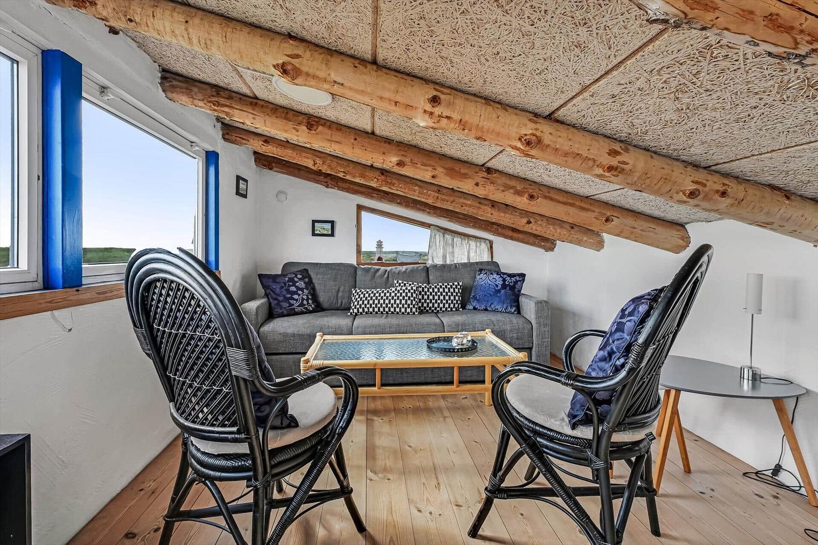 Wohnbereich unter Dachschräge mit Holzbalken, grauem Sofa und Korbsesseln.
