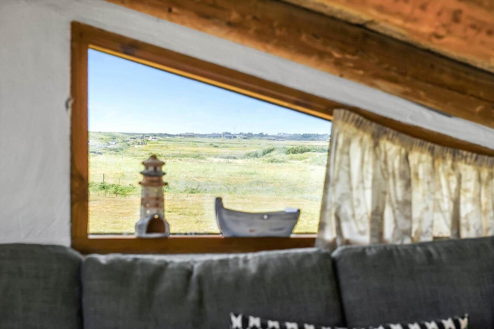 Dachfenster mit Leuchtturmfigur, Vorhang und Blick auf weite Wiesen.