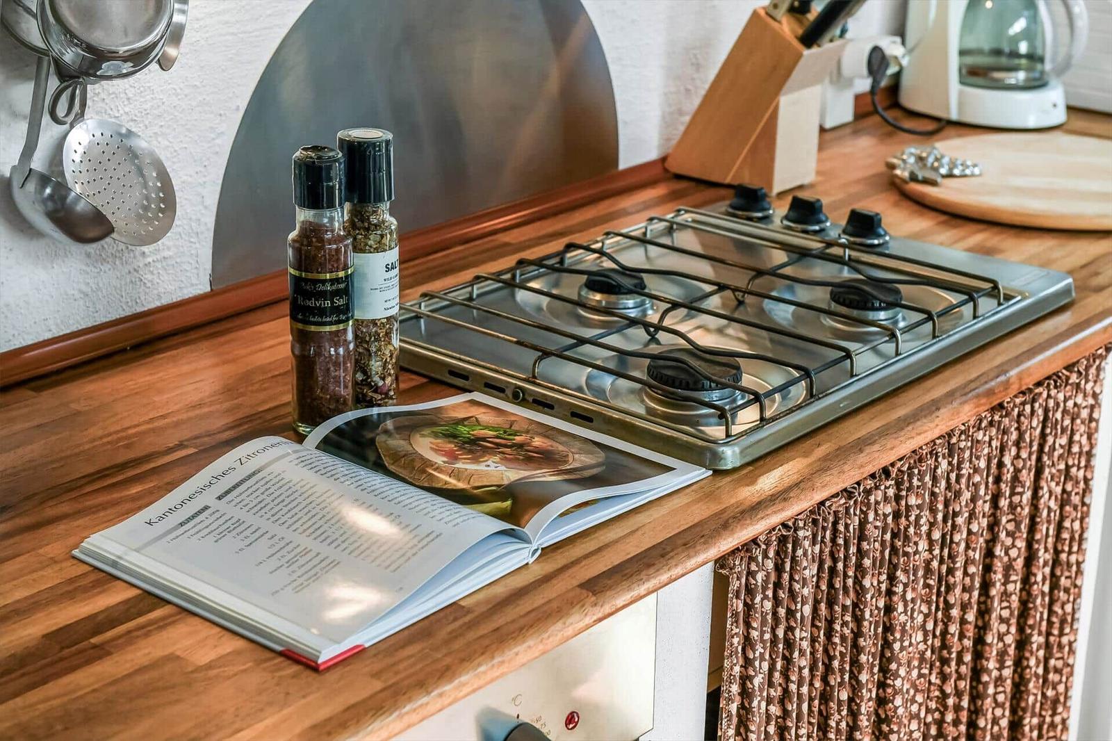 Gaskochfeld aus Edelstahl auf Holzplatte mit Kochbuch und Gewürzmühlen.