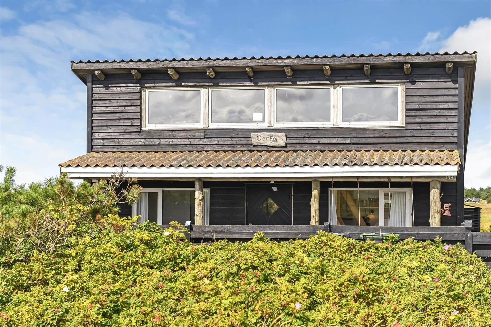 Zweistöckiges Holzhaus mit dunkler Fassade hinter einer blühenden Hecke.