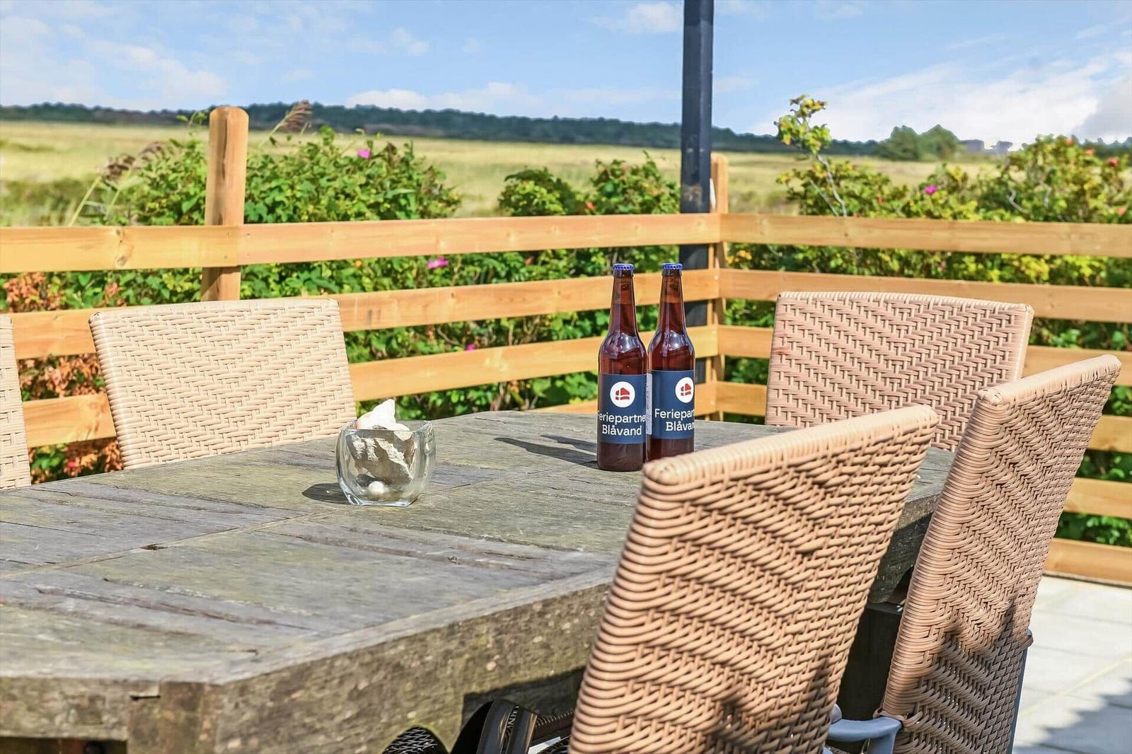 Terrasse mit Holztisch, Korbstühlen und Blick über den Zaun aufs Feld.