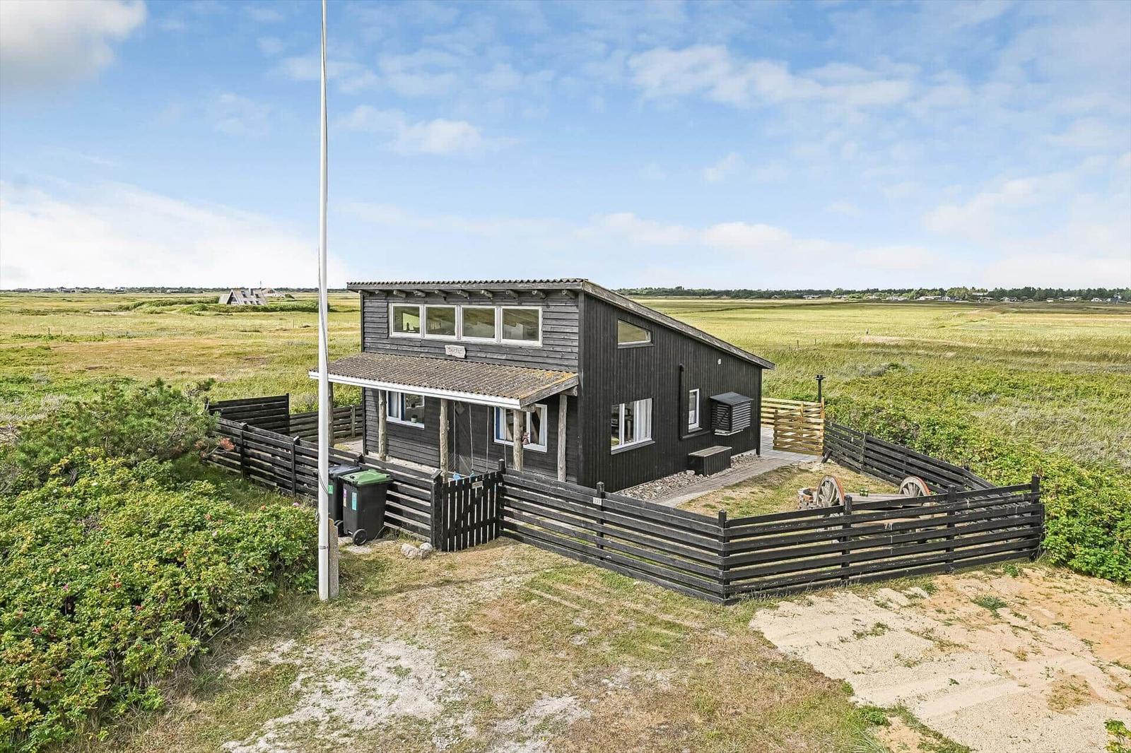 Schwarzes Holzhaus mit Zaun, Flaggenmast und Wagenrädern in offener Landschaft.