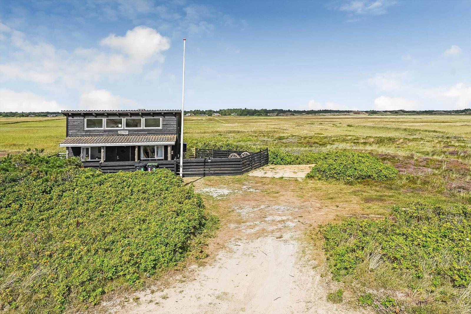 Dunkles Holzhaus mit Flaggenmast und Sandweg durch Heide und Sträucher.