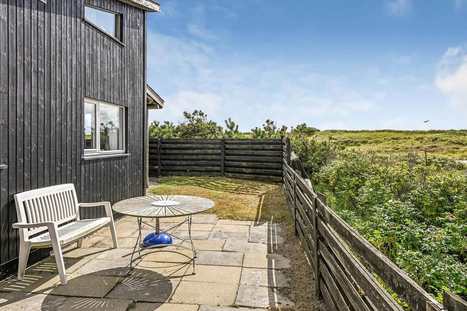 Steinterrasse mit weißer Gartenbank, rundem Holztisch und dunklem Holzzaun.