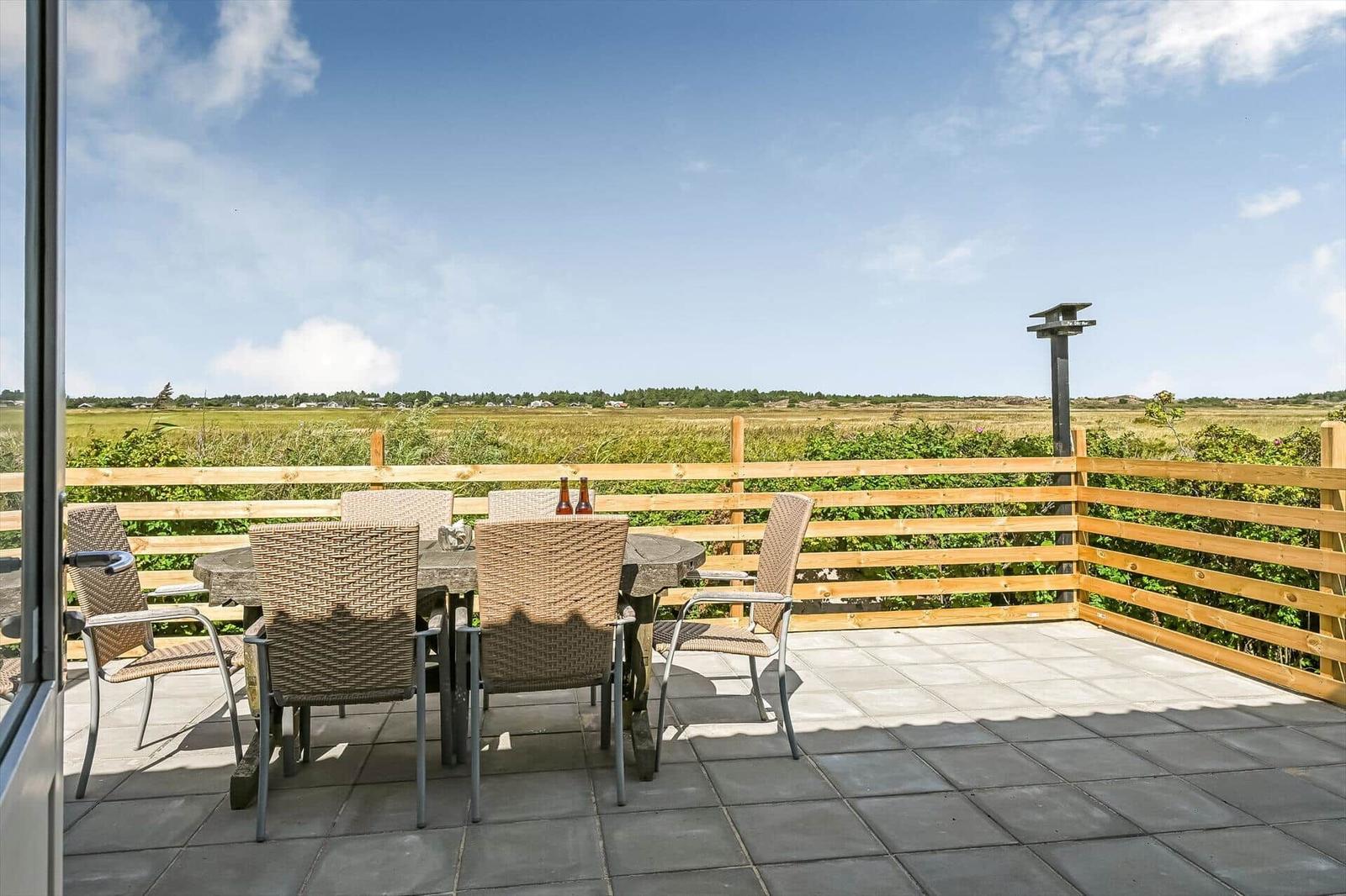 Terrasse mit Holztisch, Korbstühlen und Holzzaun vor weiter Wiesenlandschaft.