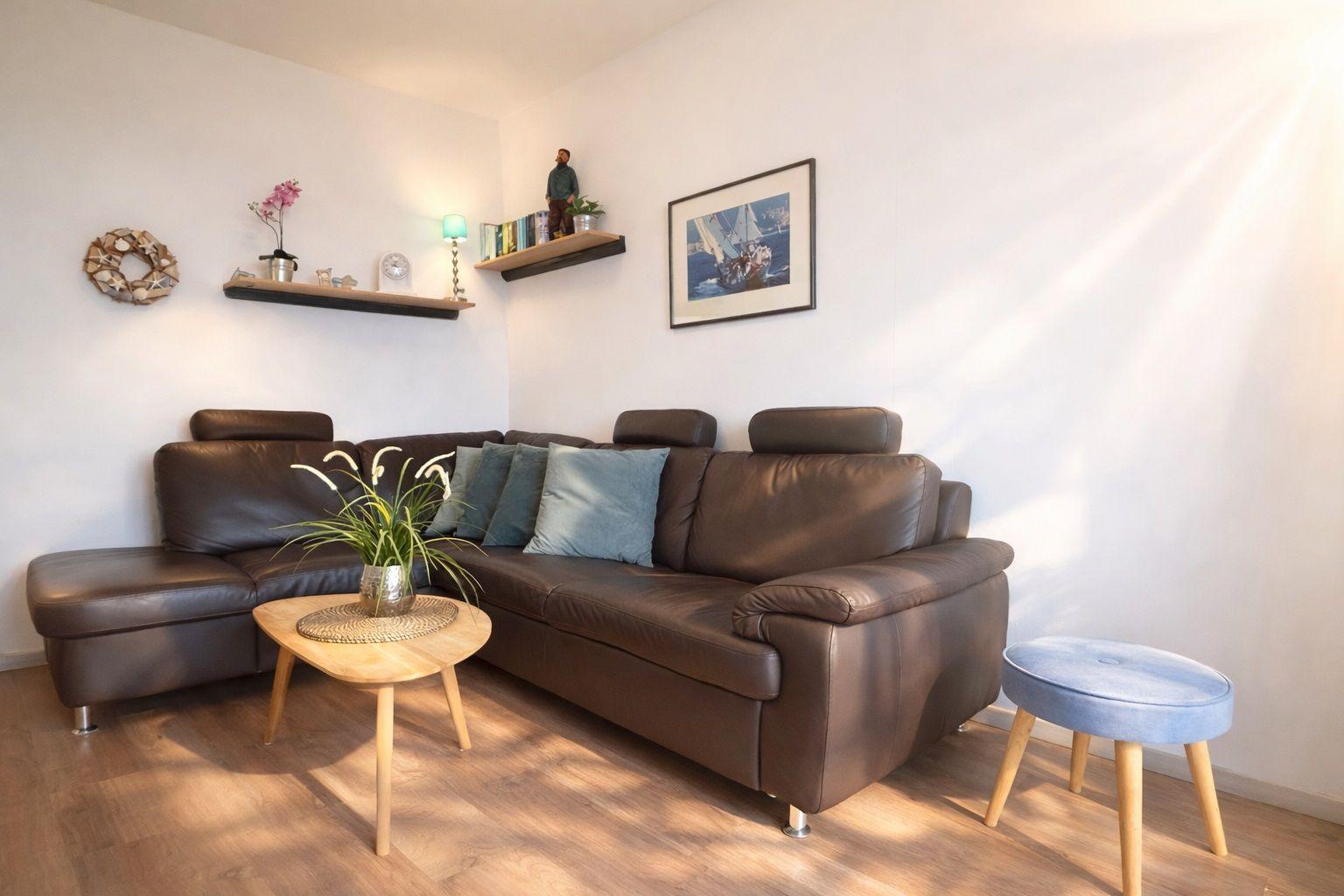Ein Wohnzimmer mit braunem Sofa, Holztisch und blauem Hocker. An den Wänden hängen Bilder und Regale.