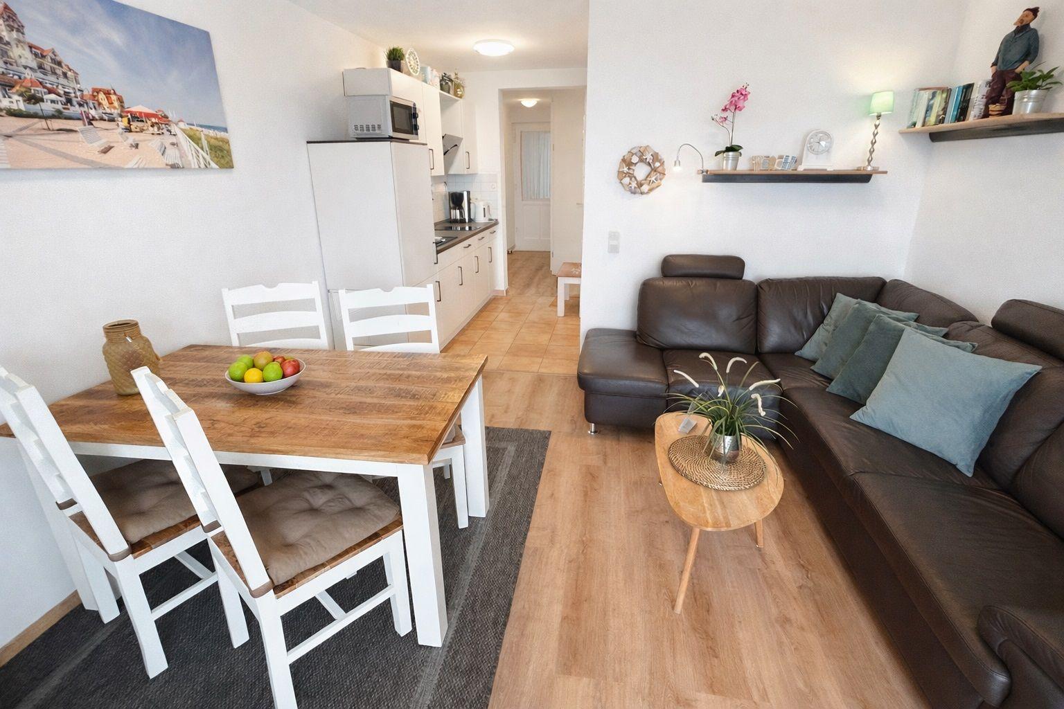 Wohn- und Essbereich mit braunem Sofa, Holztisch und weißen Stühlen. Küche im Hintergrund.