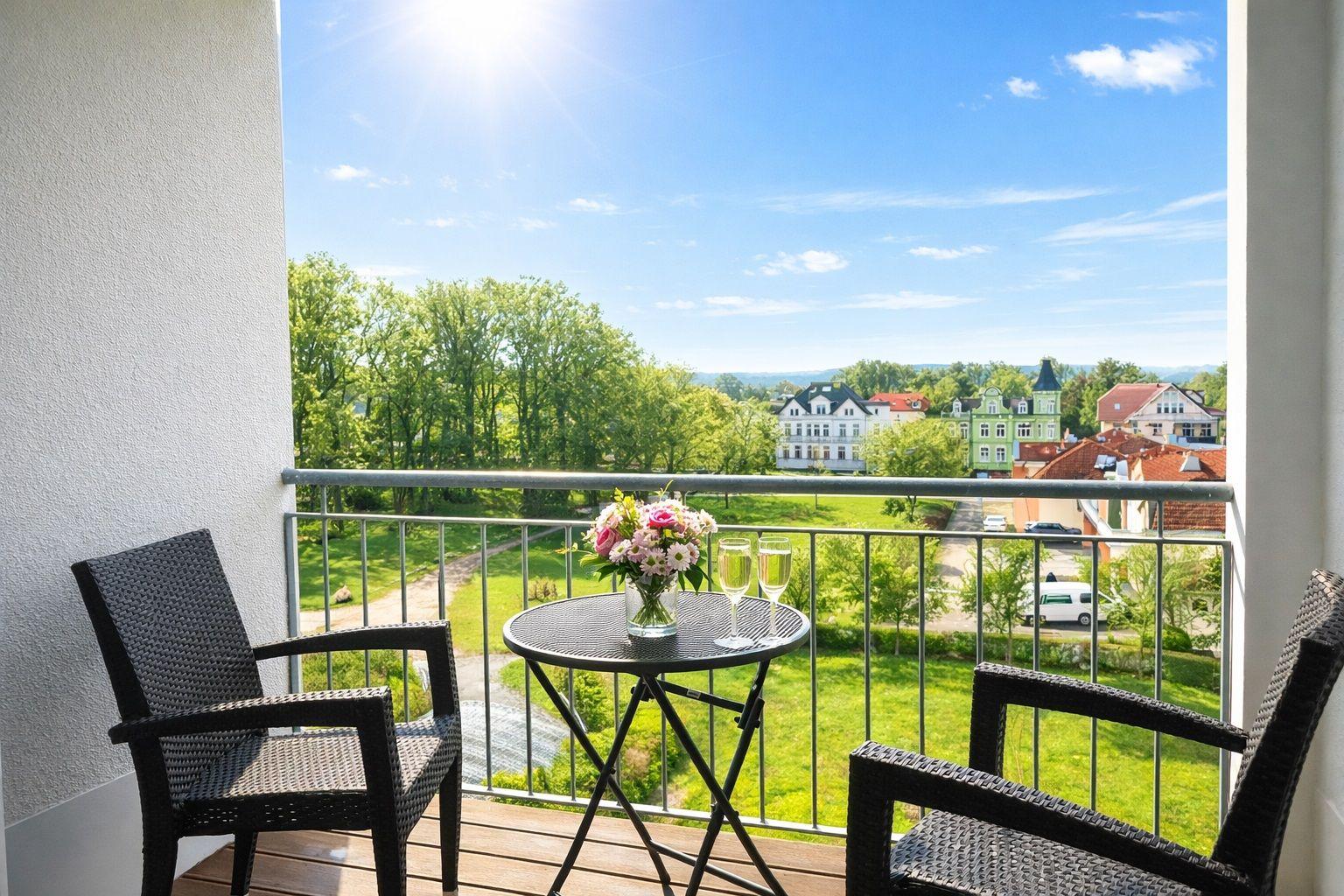 Terrasse mit Tisch, zwei Stühlen und Blumen. Blick auf grüne Landschaft und Gebäude.