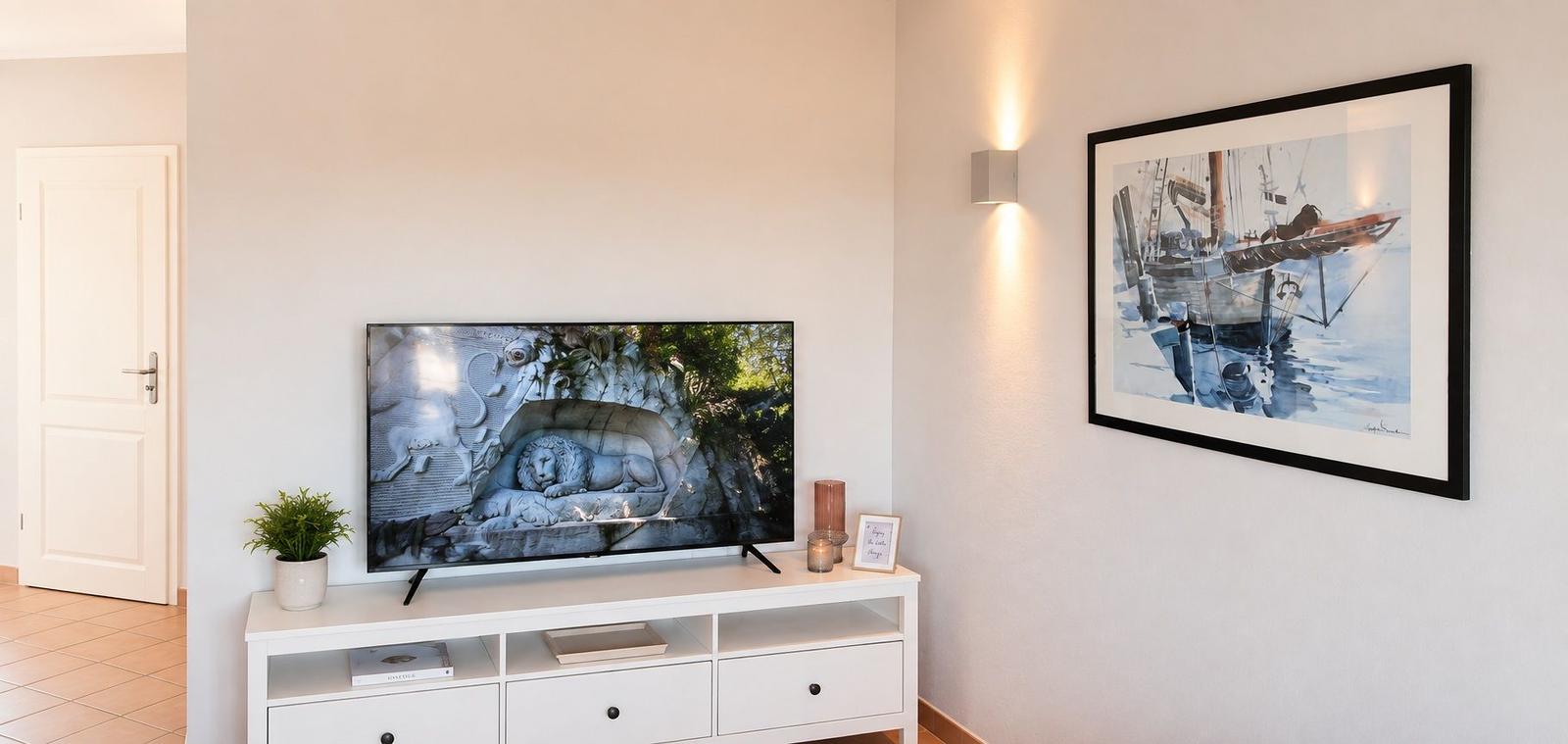 TV auf weißem Tisch, Wandbild mit Segelboot, Wandlampe und Tür im Hintergrund.