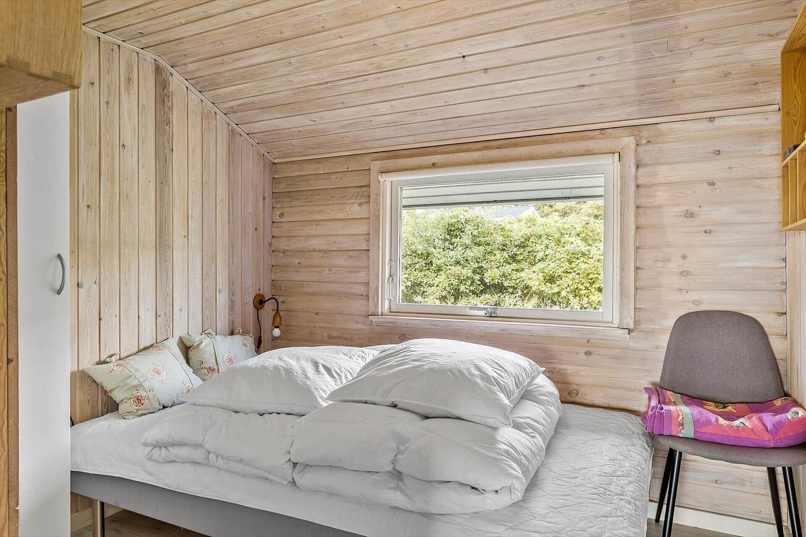 Ein Schlafzimmer mit Holzwand, Bett und Fenster. Draußen ist ein Sonnenschirm sichtbar.
