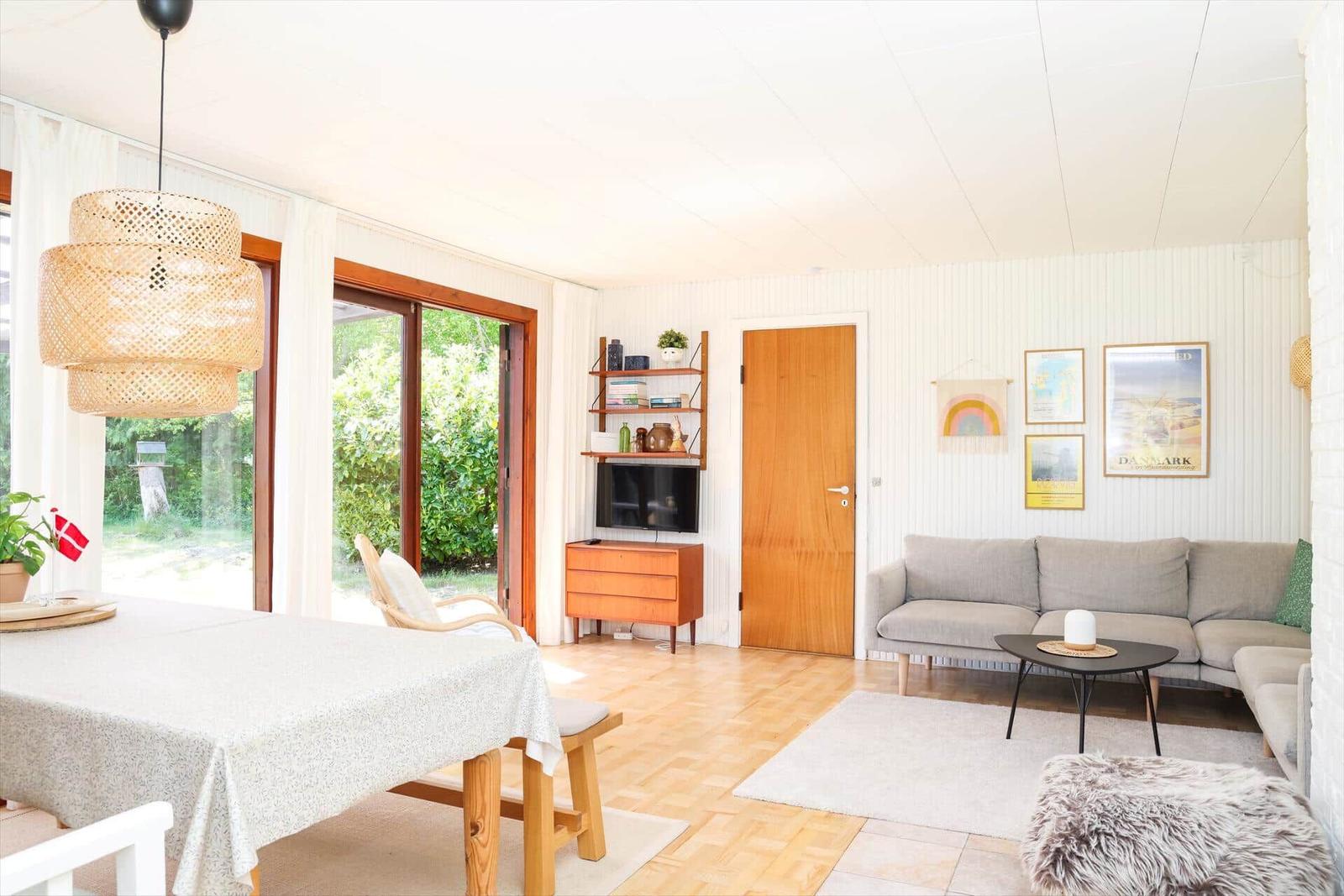 Terrasse mit Holztisch, Tischdecke und Blumen unter Glasdach. Umgeben von Pflanzen und dunkler Wand.