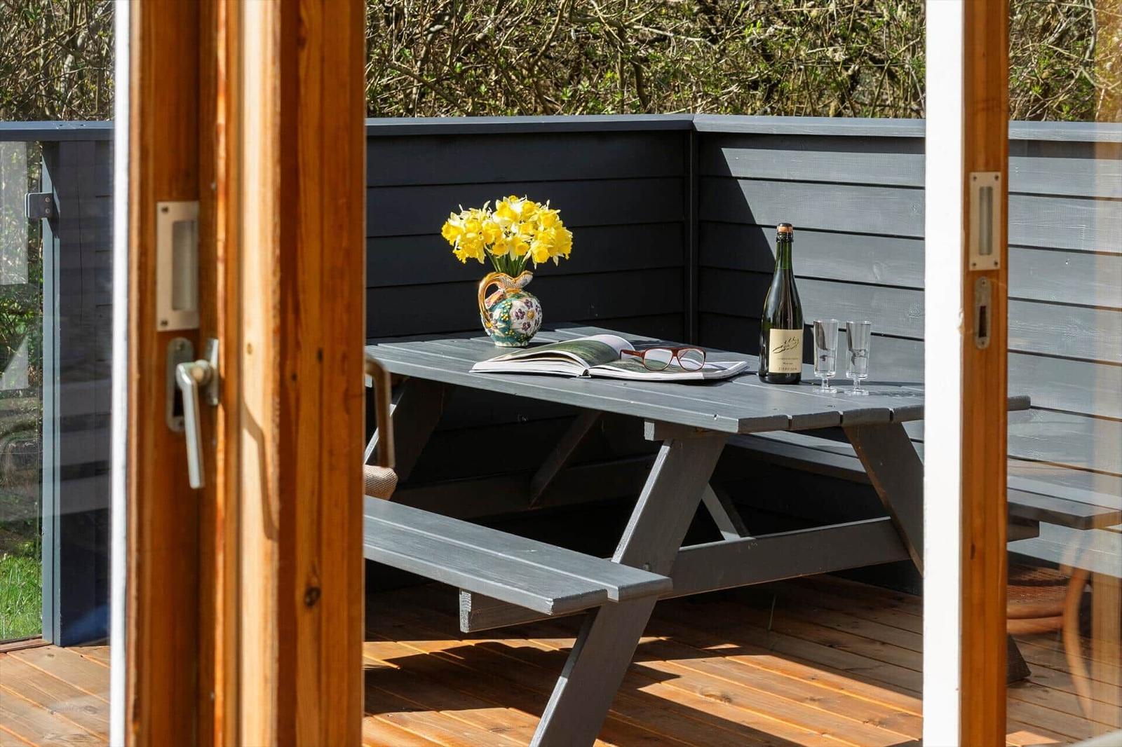 Terrasse mit Tisch, Blumen und Flasche. Blick auf grüne Bäume und Haus.