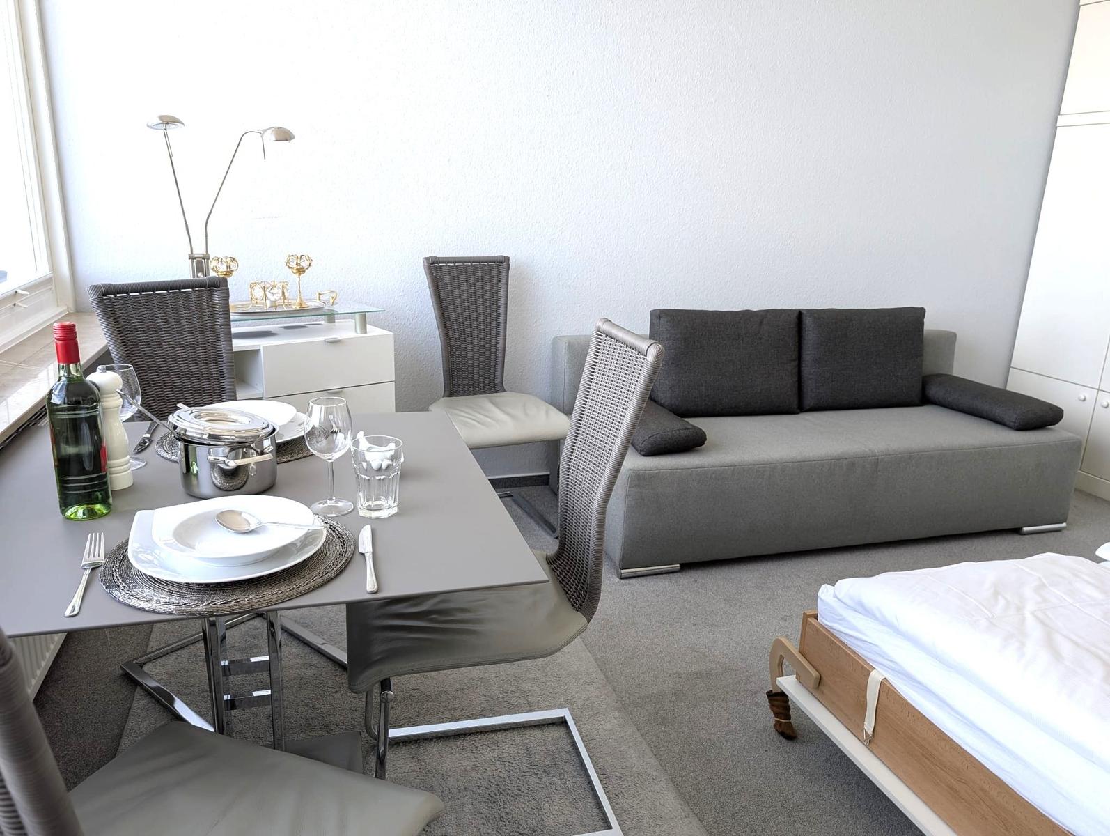 Gedeckter Esstisch, graues Sofa, weiße Kommode und Bettkante auf Teppich.