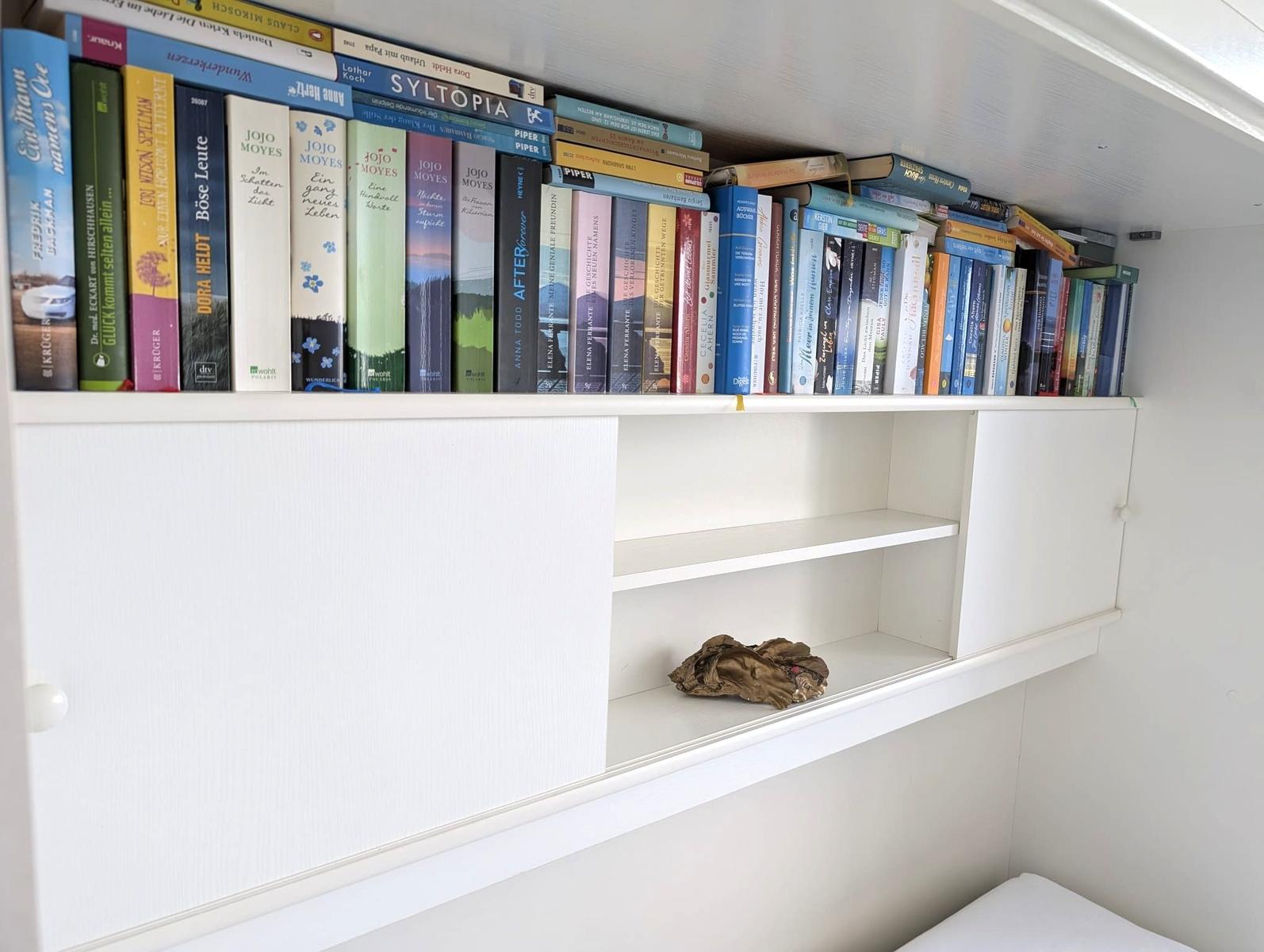 Weißes Wandregal mit Bücherreihe, Schiebetüren und offenen Fächern.