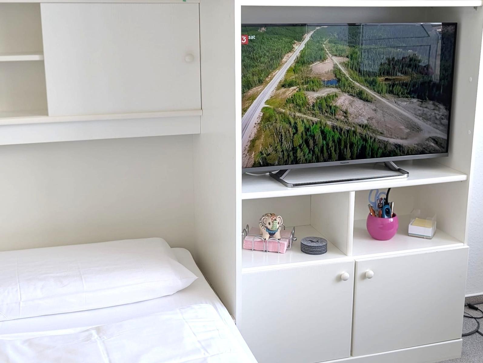 Weißes Regal mit Fernseher neben dem Bett mit weißer Bettwäsche.