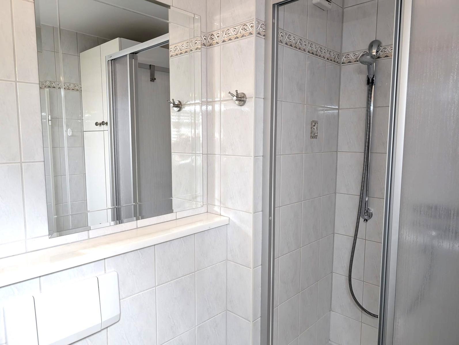 Badezimmer mit Glasdusche, großem Spiegel, Haken und weißen Fliesen.