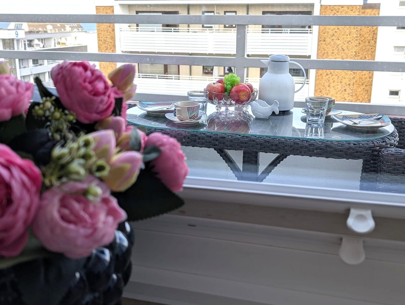 Balkontisch mit Obstschale, Kaffeegedeck und rosa Blumen vor Häuserzeile.