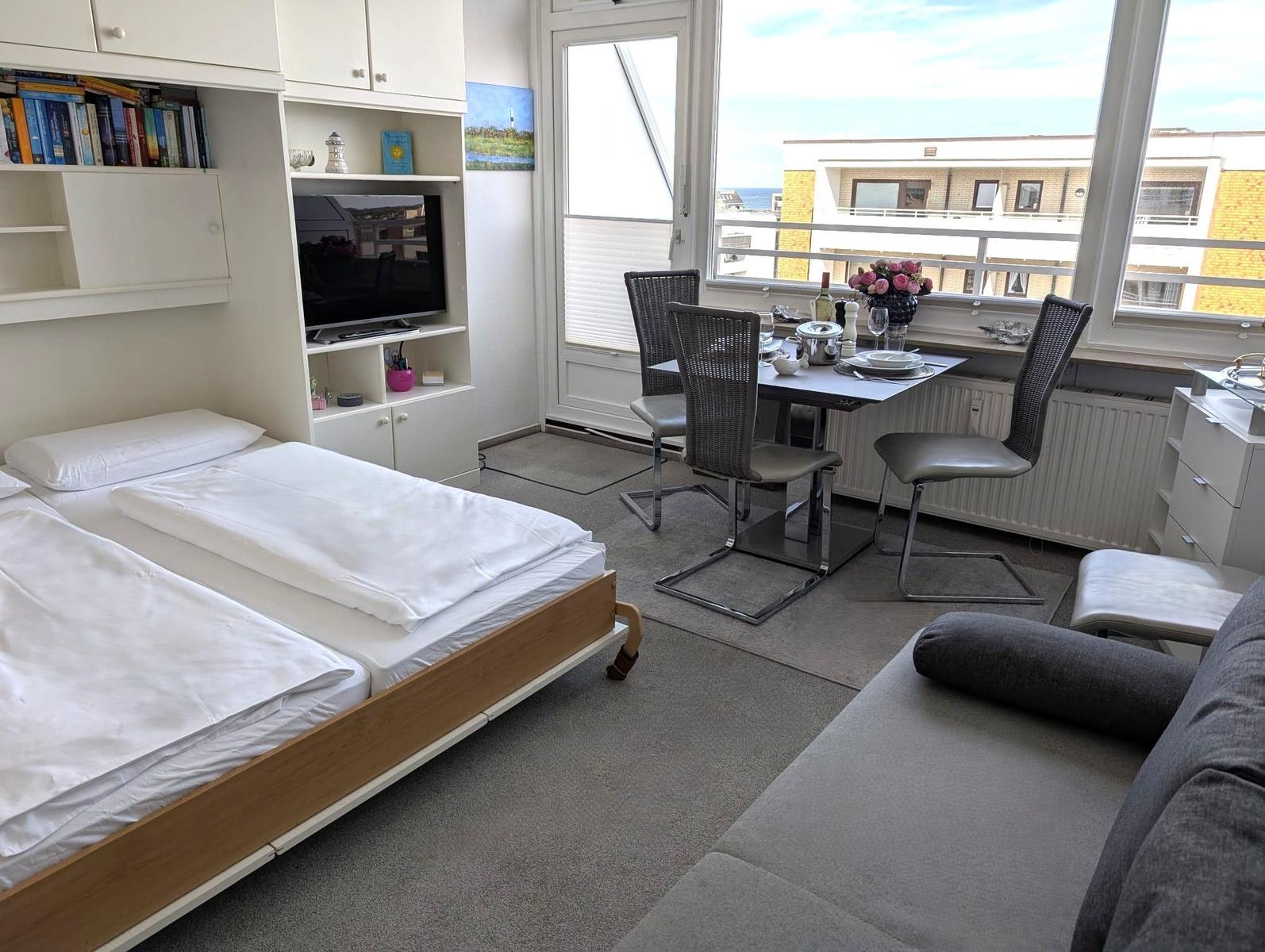 Bett mit weißer Wäsche, gedeckter Esstisch, TV-Schrank und große Fensterfront.