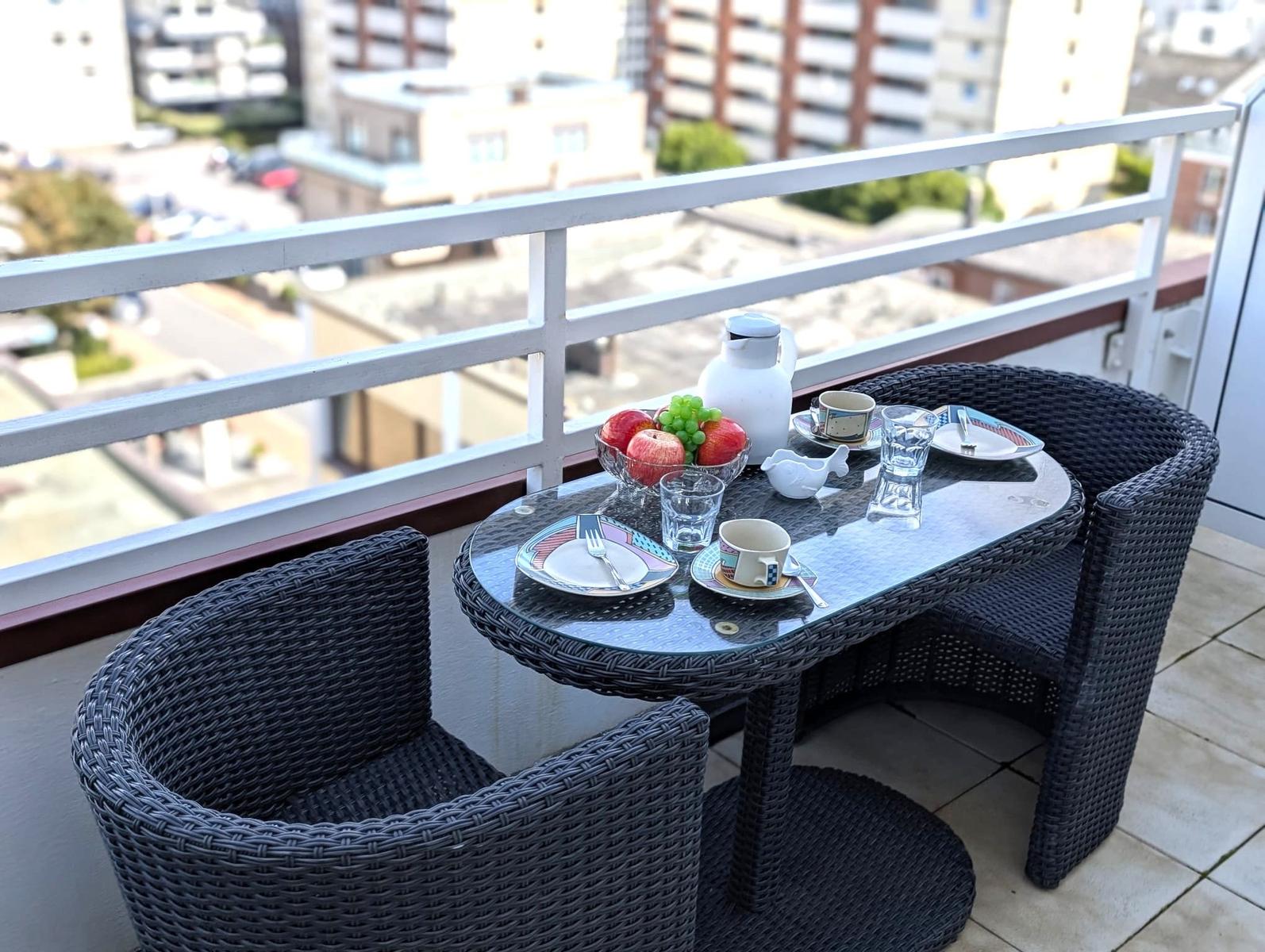 Balkon mit zwei Rattansesseln, gedecktem Glastisch und Blick auf Häuser.