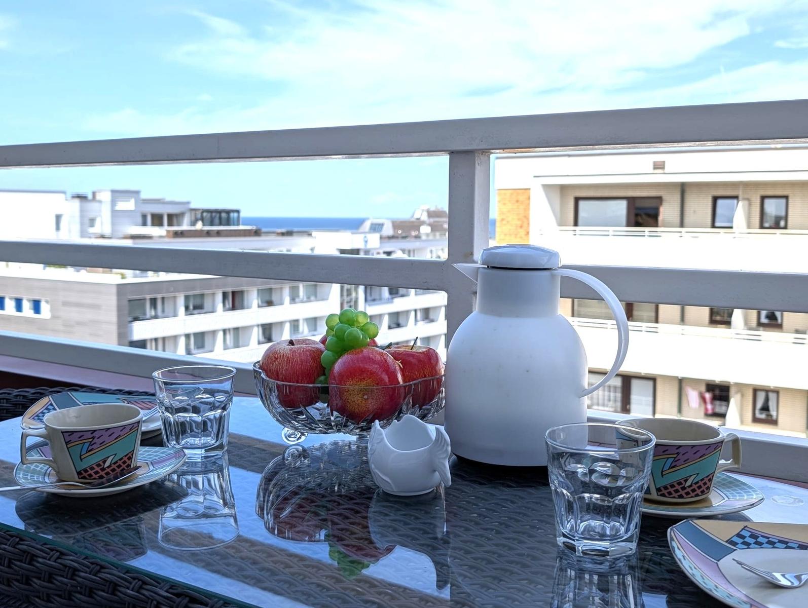 Balkontisch mit Kaffeegedeck, Obstschale und Blick auf Häuser und Meer.