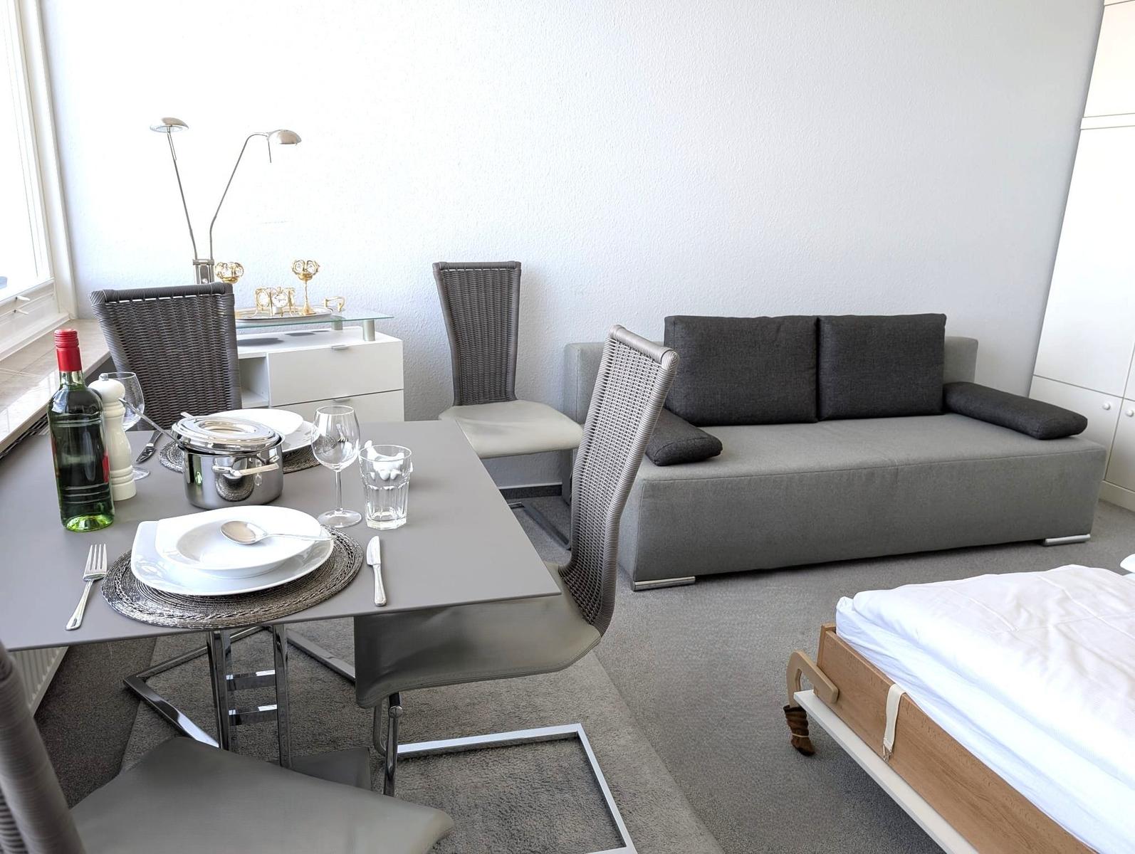 Esstisch mit Gedeck, graues Sofa und Bettkante im hellen Wohnraum.