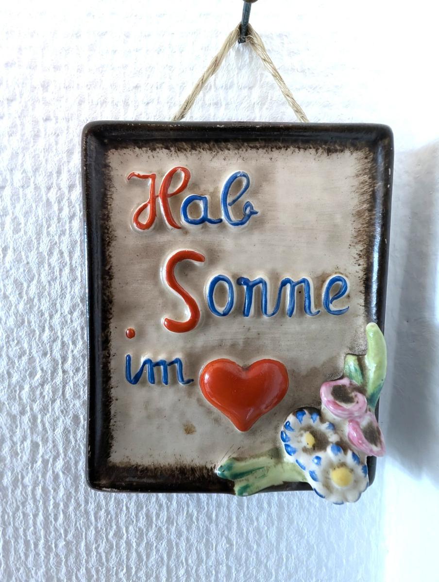 Keramikschild mit Spruch, rotem Herz und Blumenrelief an weißer Wand.