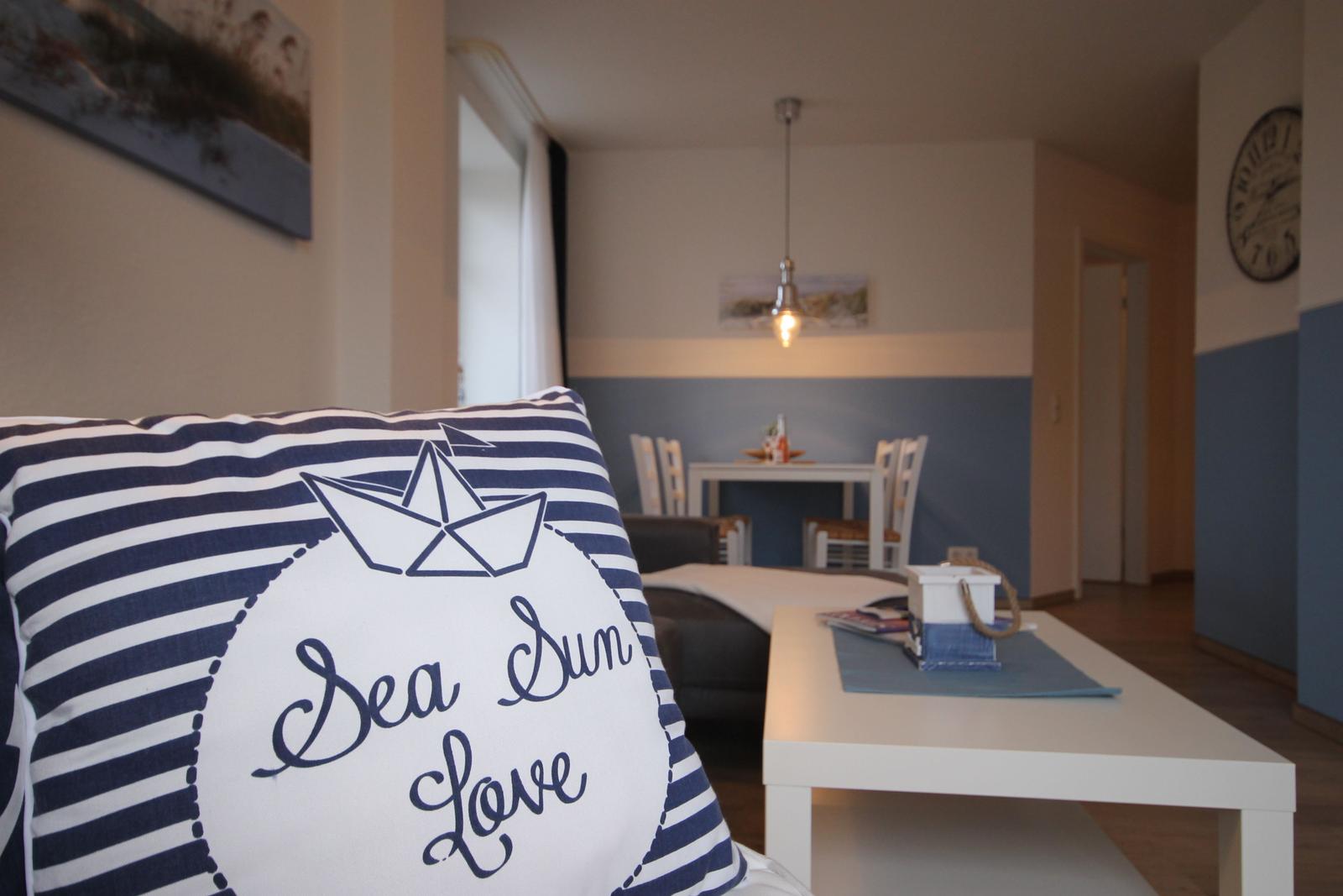 Wohnbereich mit Kissen, Tisch und Stühlen. Wandfarbe blau-weiß. Wanduhr und Bild an der Wand.
